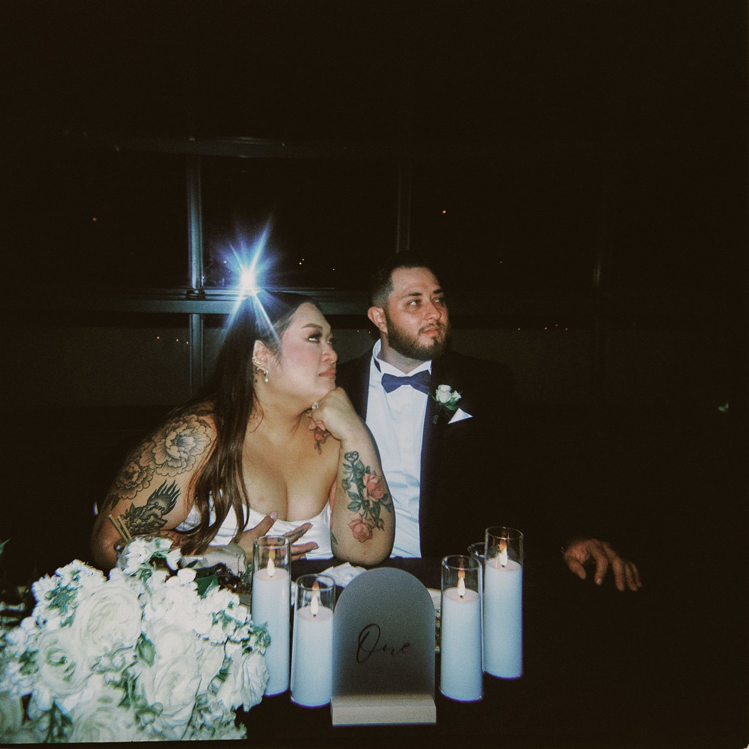 A couple sits at their reception table with candles and floral arrangements while sharing a candid moment together.