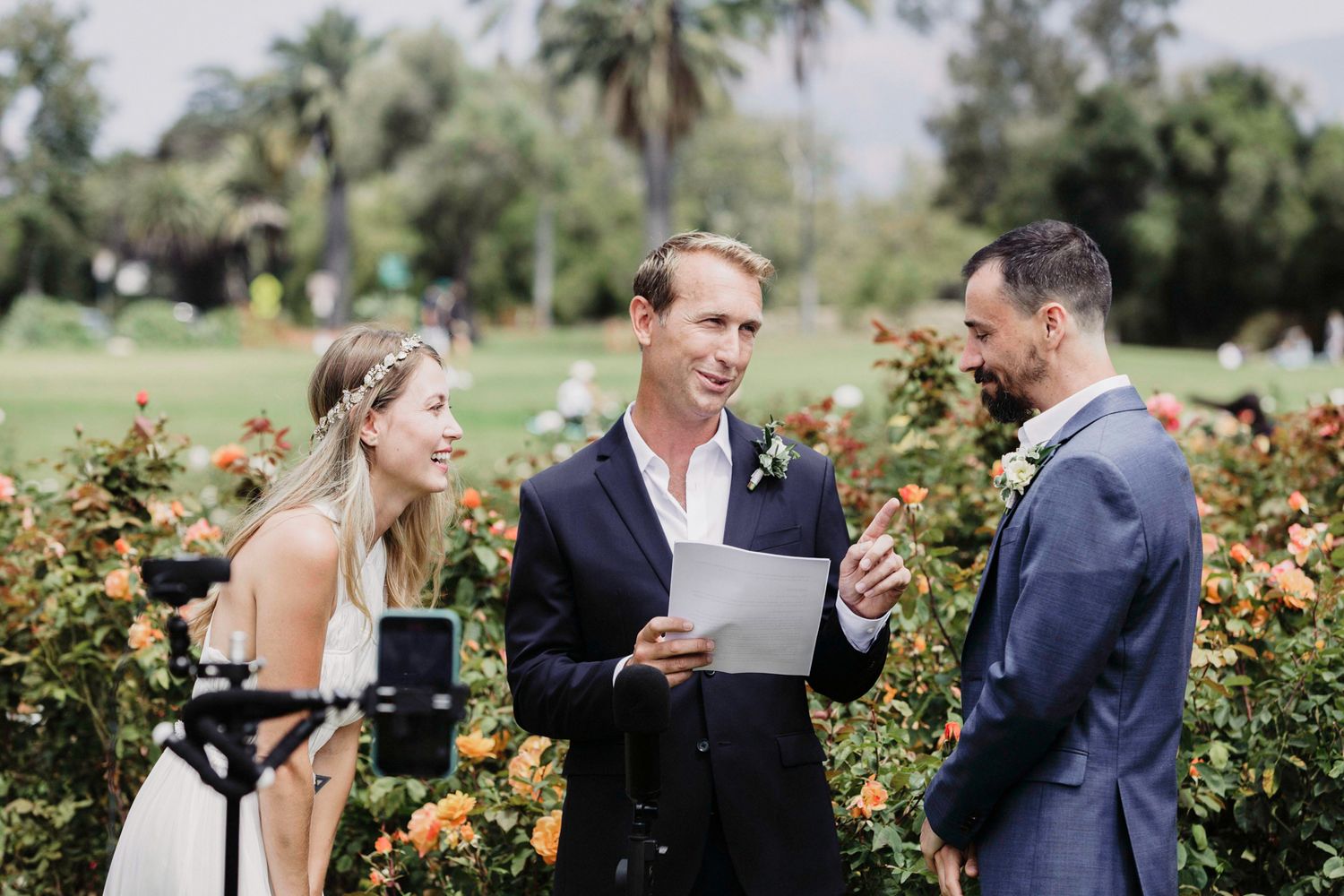 A wedding party shares joyful moments together in a lush garden setting with blooming roses and palm trees.