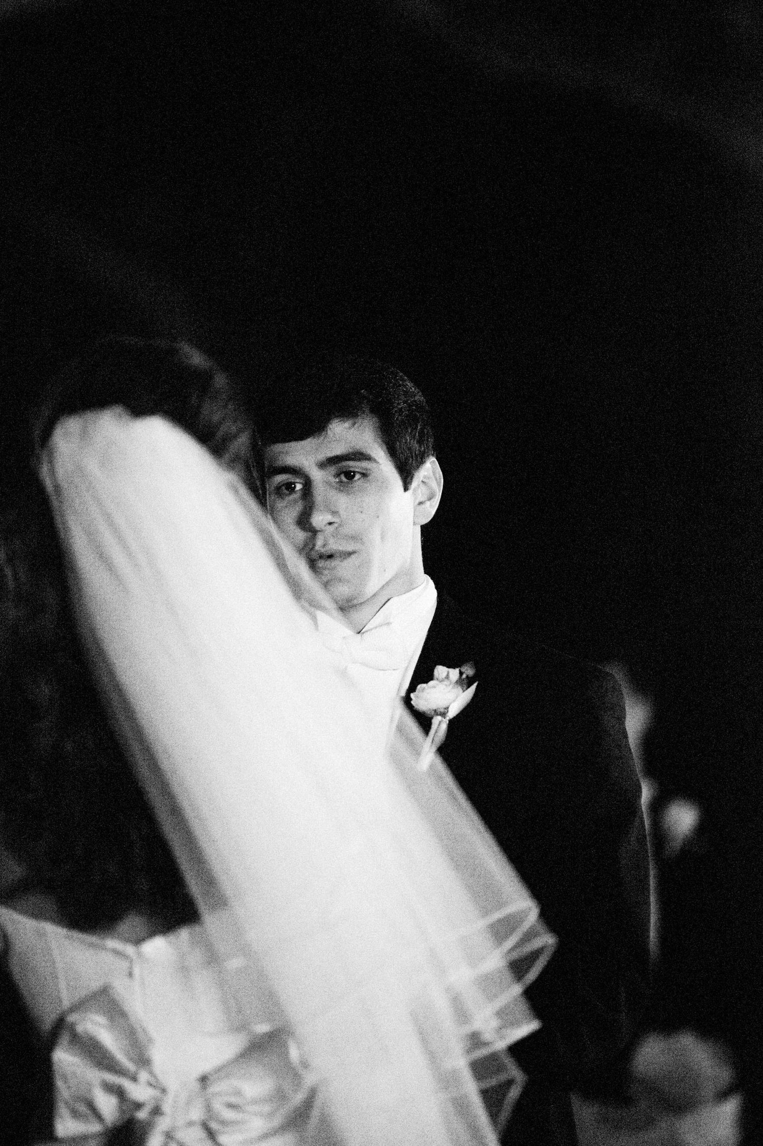 Black and white wedding photo of groom in tuxedo gazing at bride in veil during first dance.