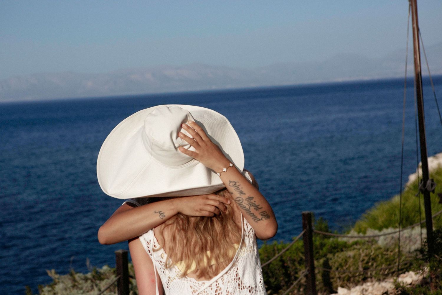 Woman in white wide-brim hat and lace dress overlooking a stunning blue Mediterranean sea on a sunny day.