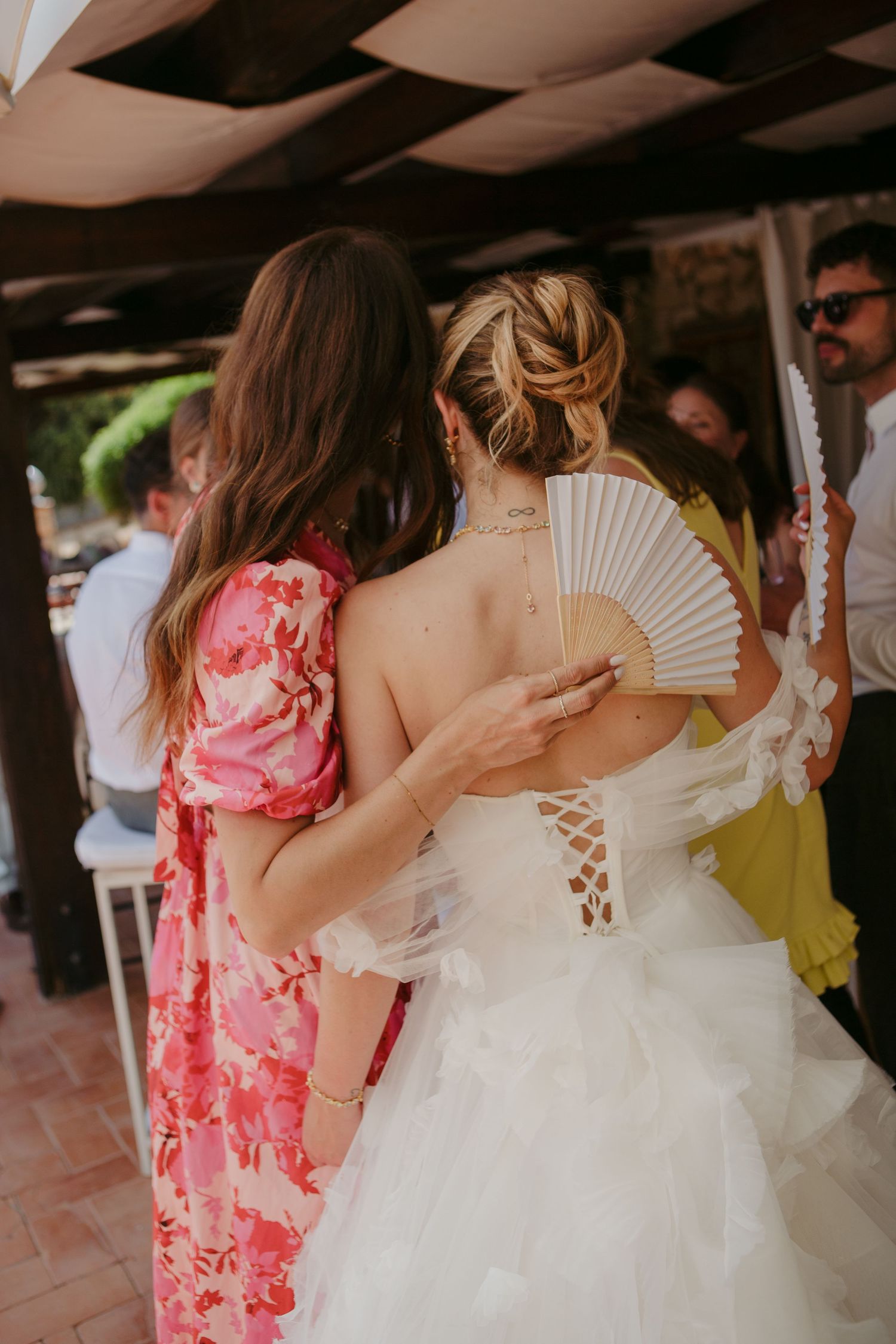 A wedding guest in a pink floral dress stands next to a bride holding a white folding fan at an outdoor reception.