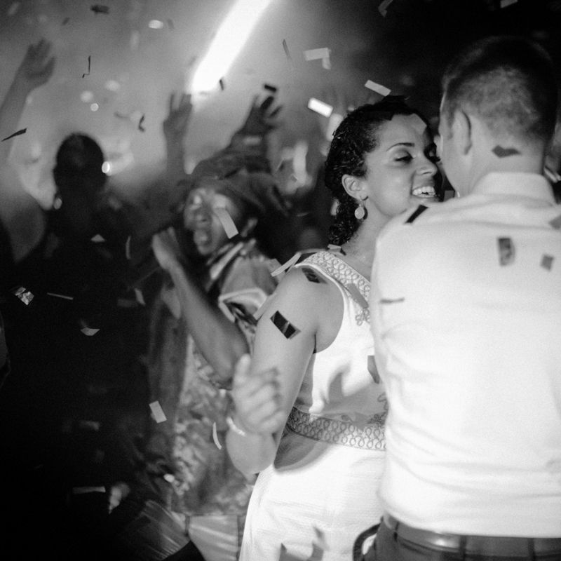 Black and white photo of couple dancing at lively party with confetti falling and crowd celebrating in background.