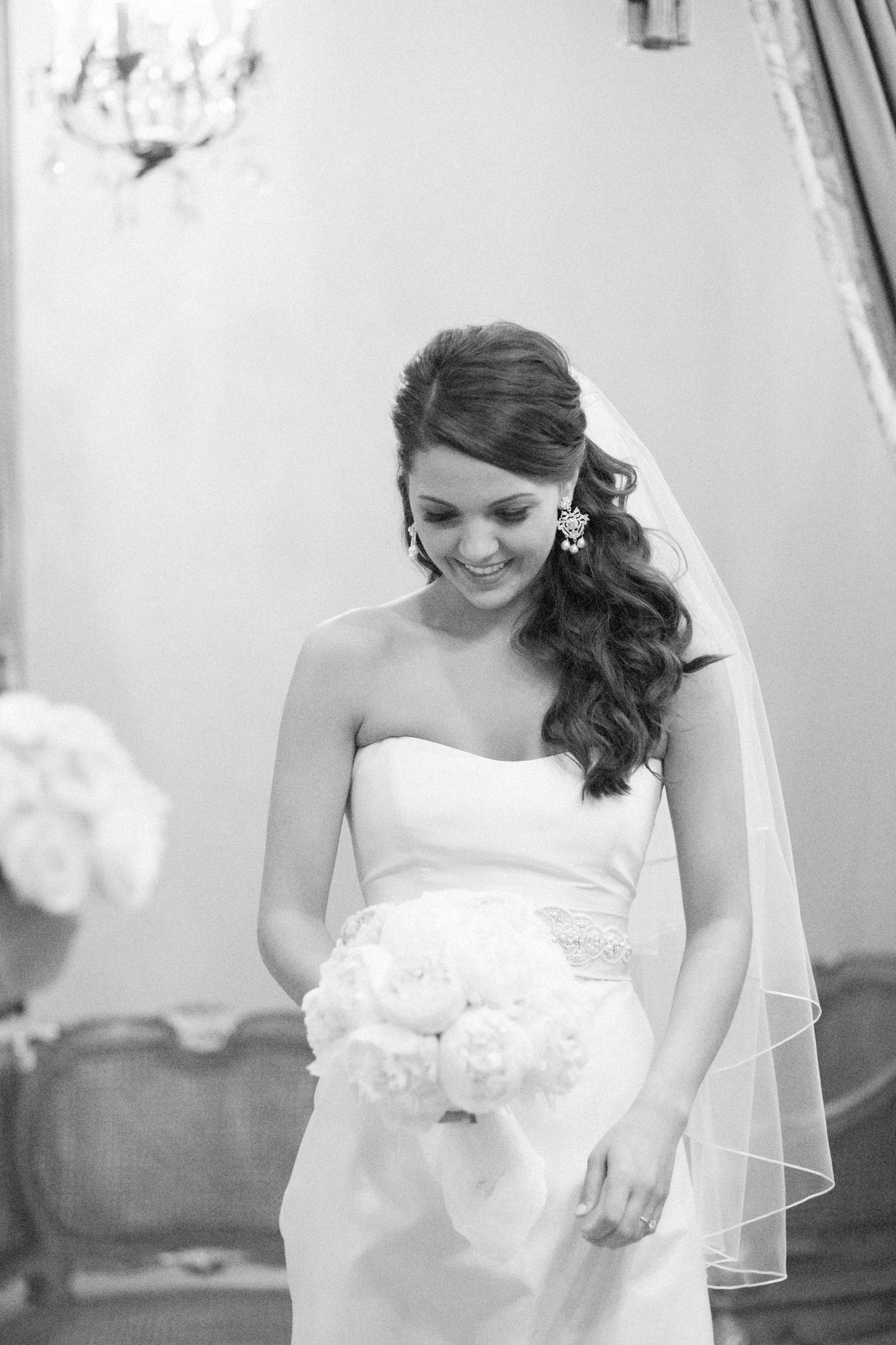 Smiling bride in strapless gown holding white bouquet with veil and chandelier earrings in black and white photo.
