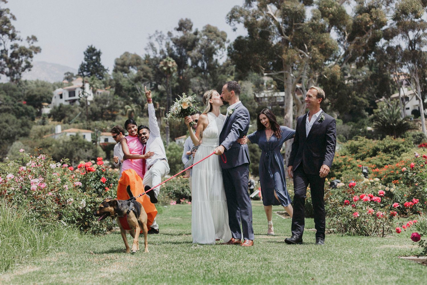Wedding party celebrates with joyful dance moves in a lush garden setting with blooming roses and trees.
