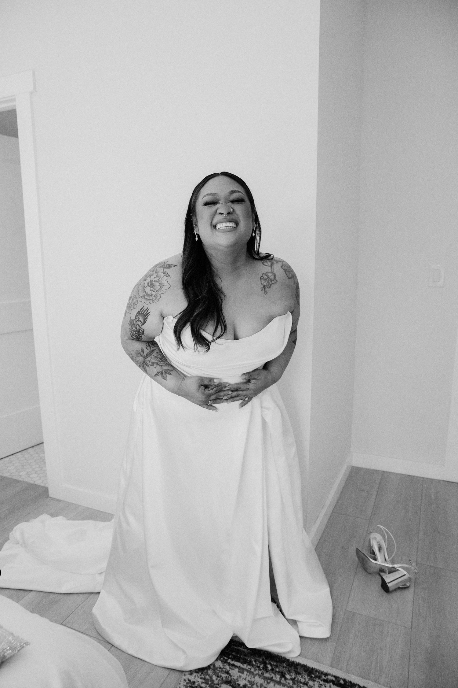 A bride in a white wedding dress laughs joyfully while showing off her tattoos in a black and white photo.