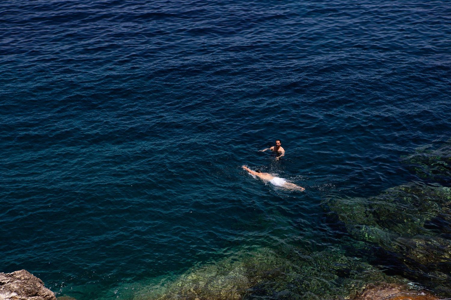 Aerial view of crystal clear turquoise ocean waters with rocky coastline and distant swimmers enjoying the sea.