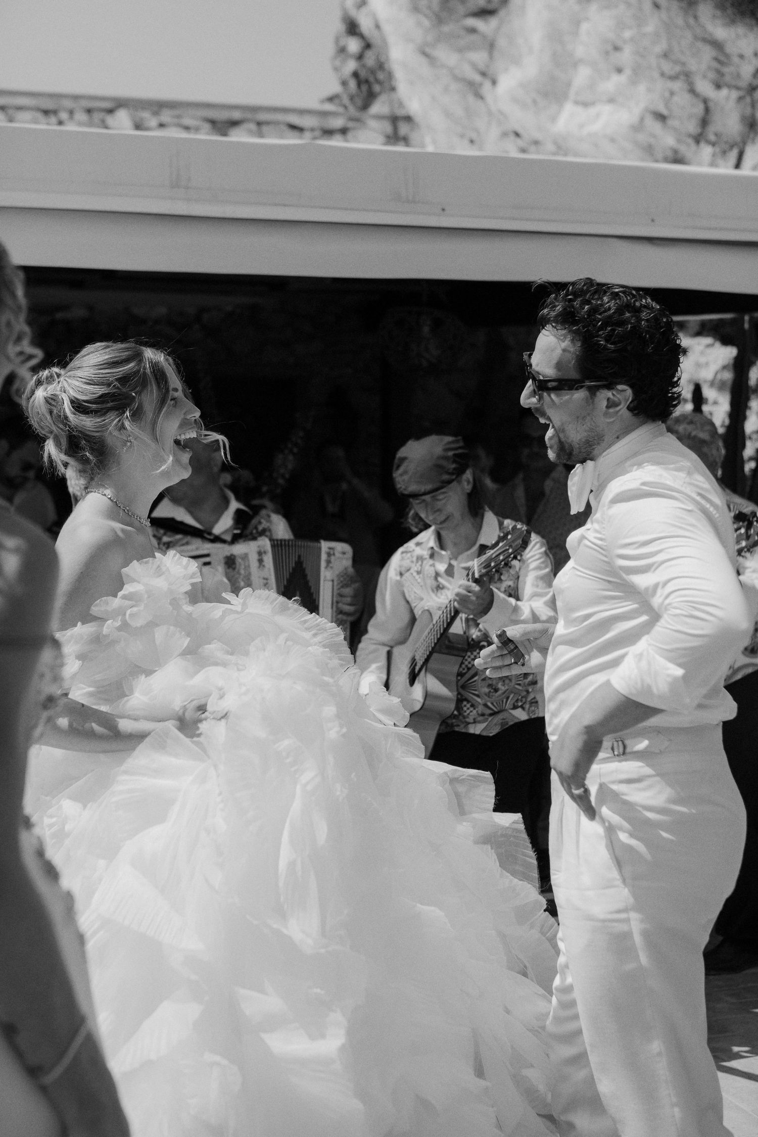 Black and white photo of a bride in a ruffled gown and groom in white suit laughing together at an outdoor wedding.