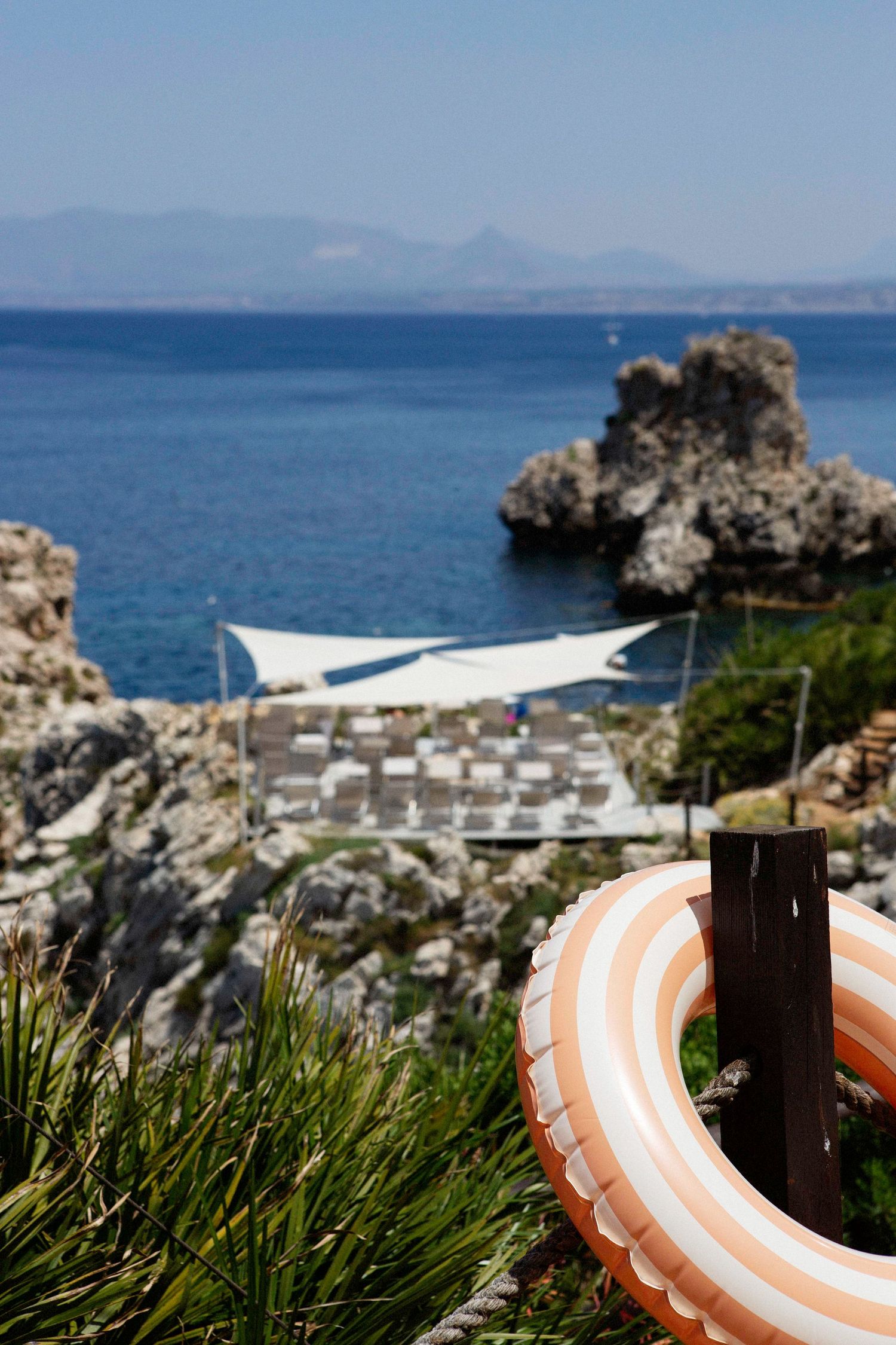 Striped inflatable ring on rocky Mediterranean coastline with beach loungers and white sail shades below.