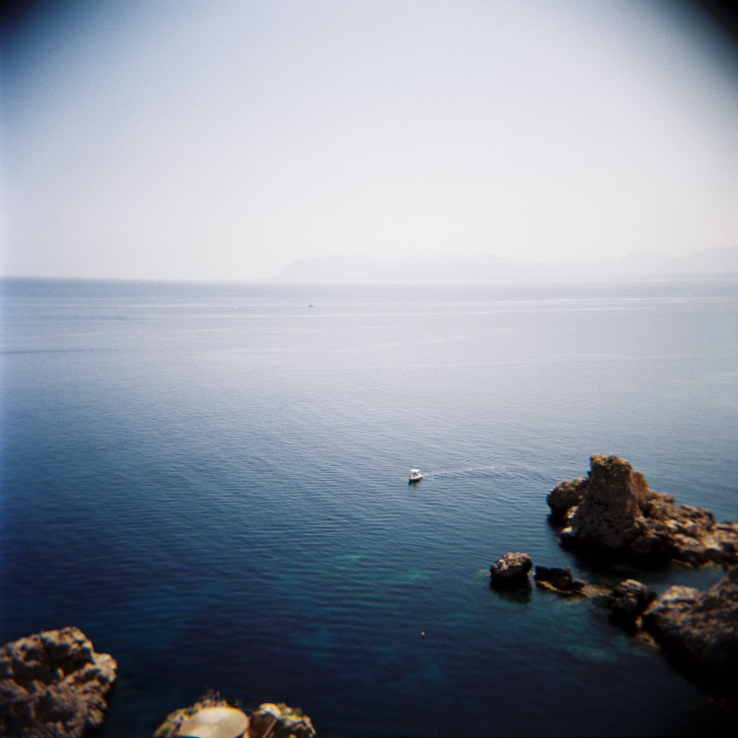 A lone boat floats on calm blue Mediterranean waters near rocky coastline with misty mountains in the distance.
