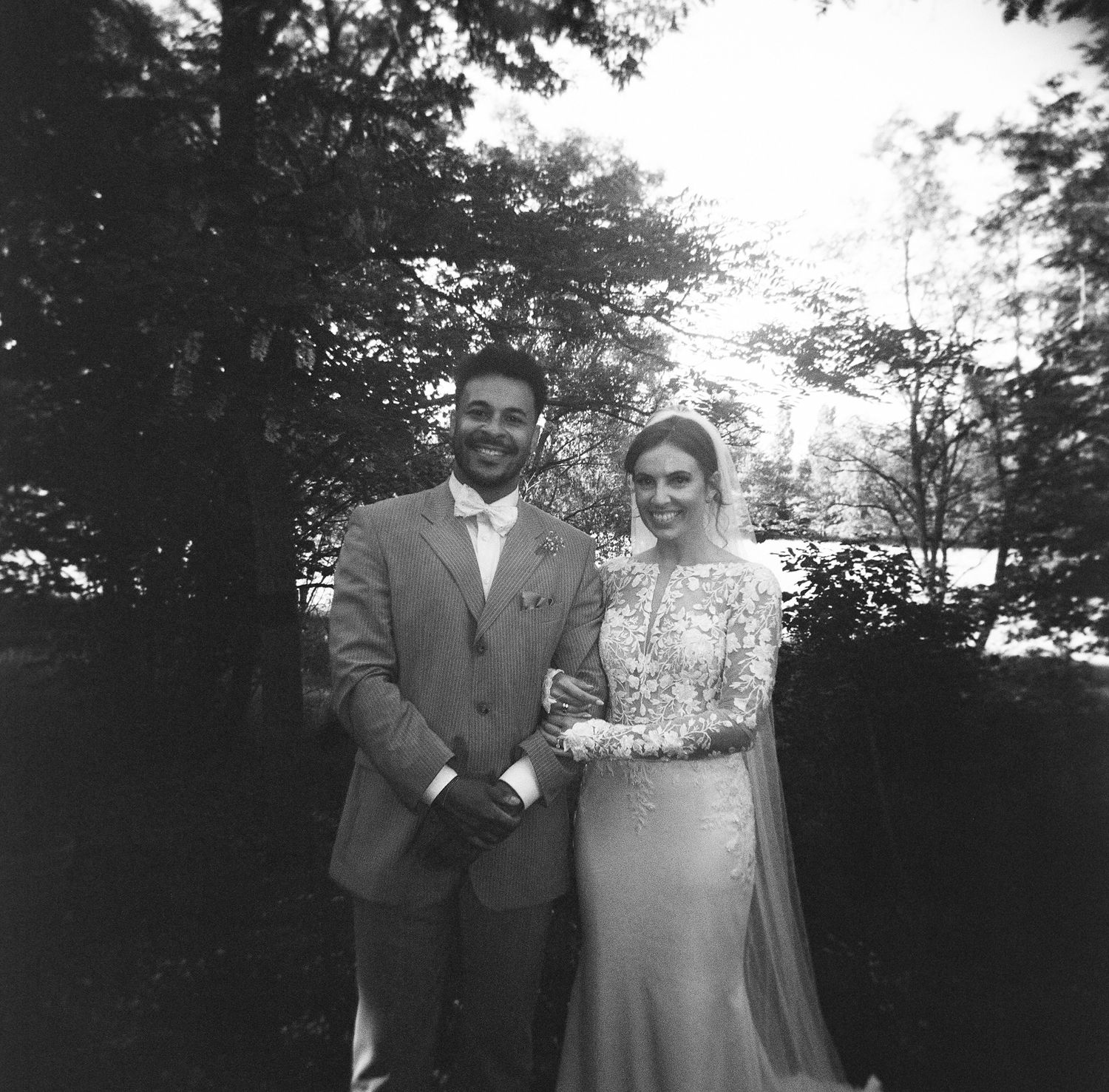 A couple poses together outdoors in black and white vintage wedding attire with trees in the background.