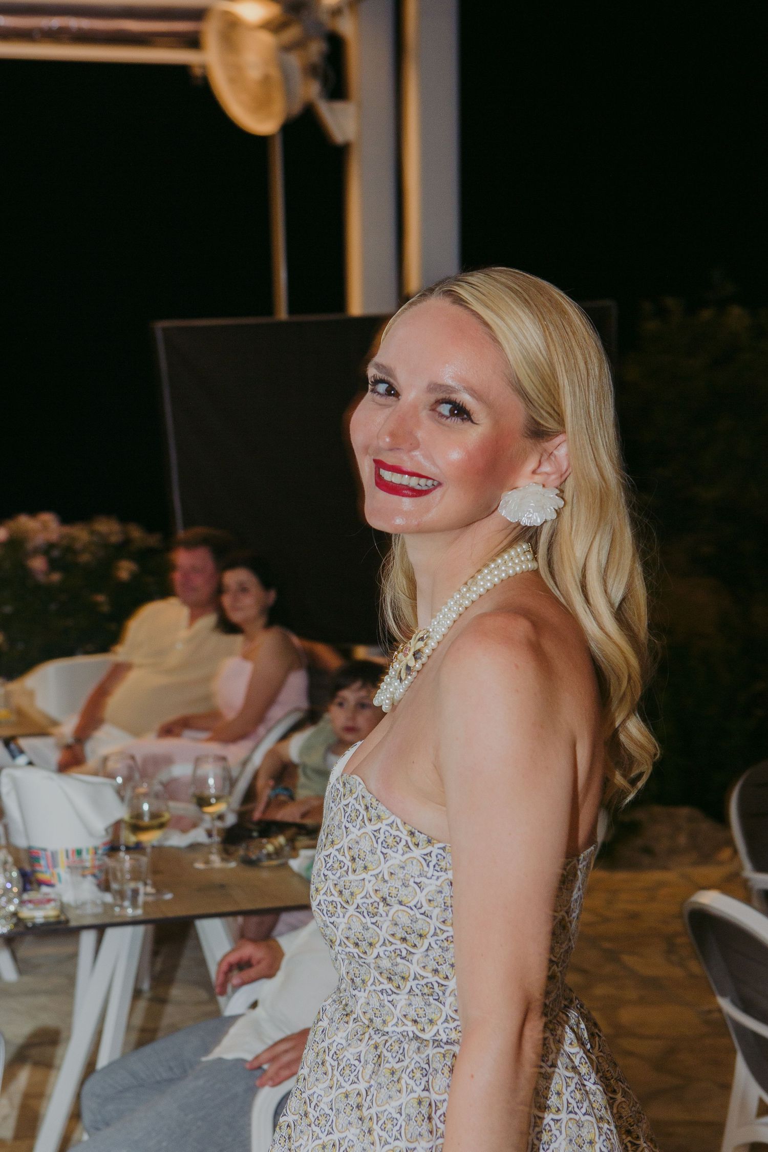 A bride in a beaded white gown and pearl necklace smiles at an outdoor wedding reception.