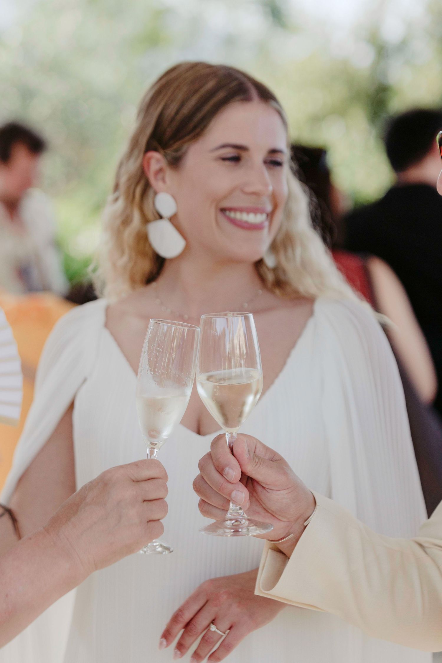 A person in a white dress toasts with champagne glasses at an outdoor celebration while wearing statement earrings.