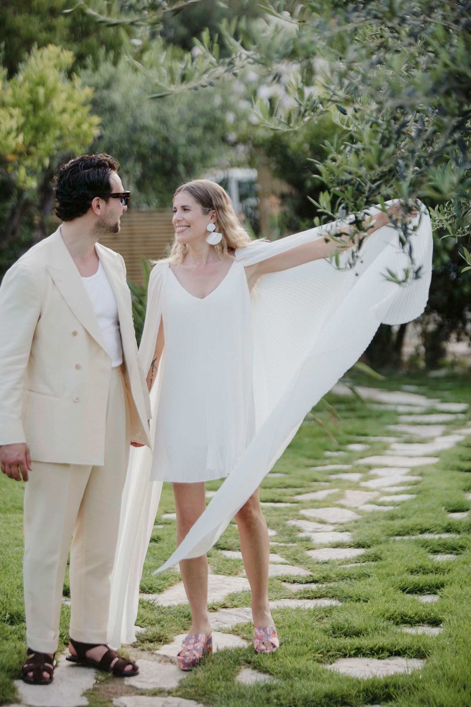 A couple in light colored outfits stands together on a garden path surrounded by greenery on a sunny day.