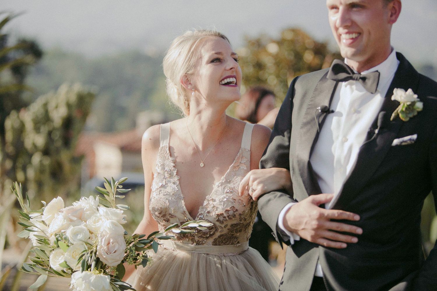 Laughing bride in floral gown holding white bouquet walks arm-in-arm with groom in black tuxedo outdoors.