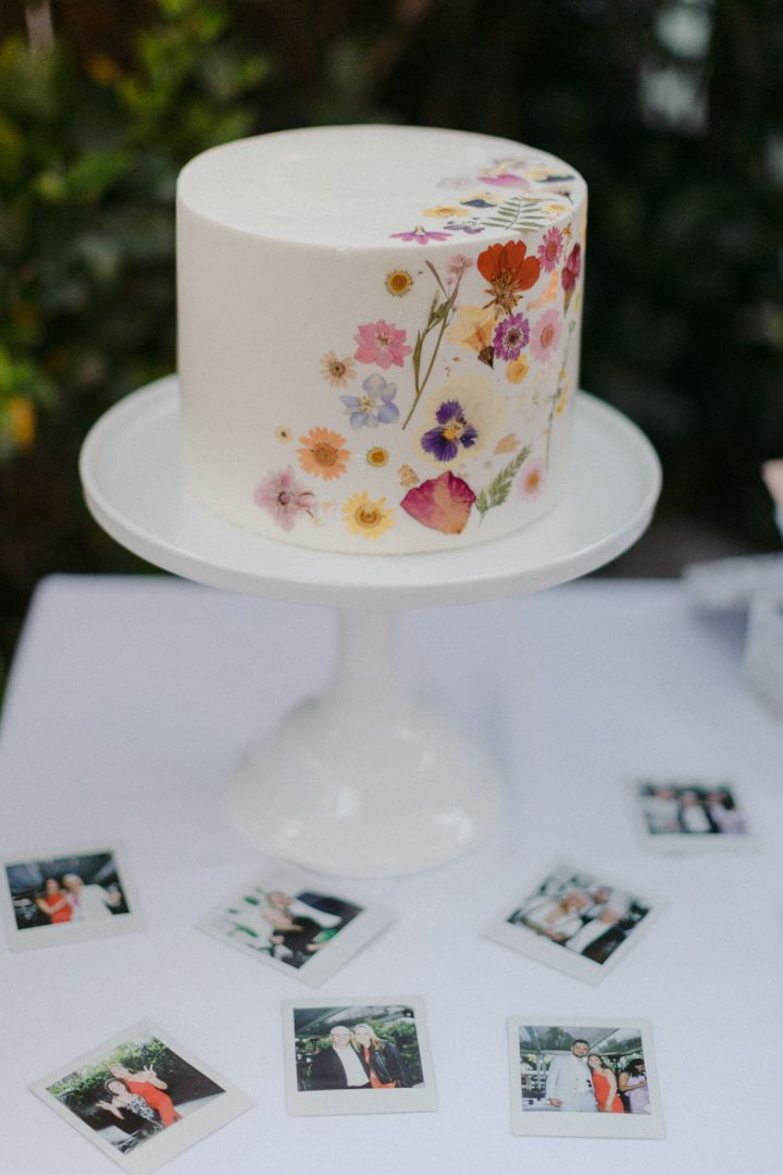 A white wedding cake decorated with delicate pressed wildflowers sits on a table surrounded by scattered polaroid photos.