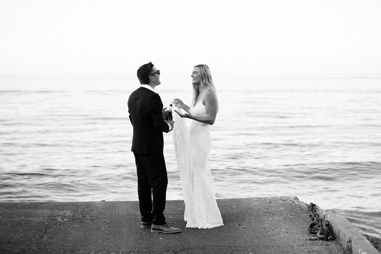 A couple in formal attire walks along a beach at sunset holding hands with ocean waves in the background.