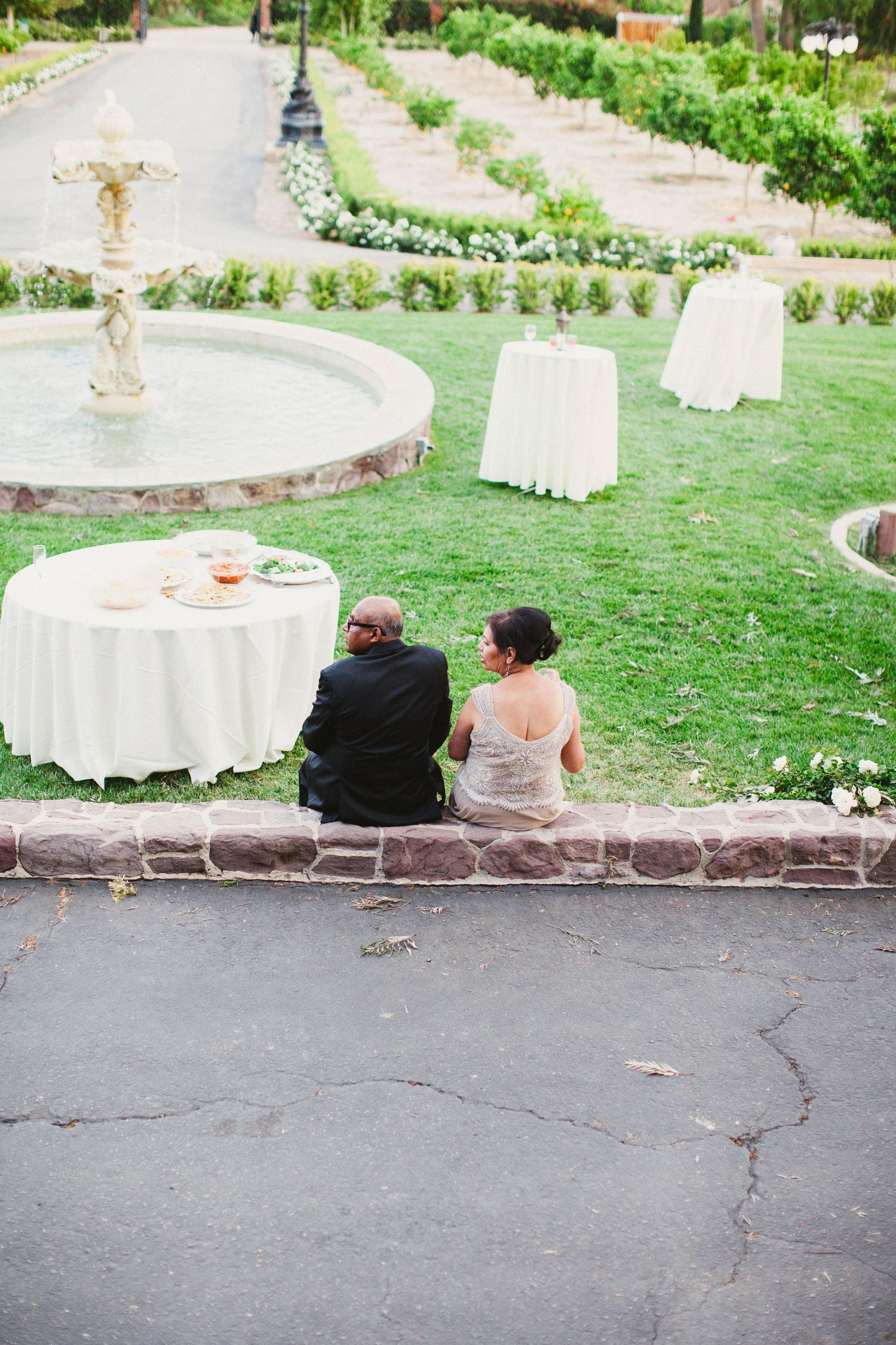Two wedding guests sit together on a stone wall overlooking an elegant outdoor reception with white tables and decor.