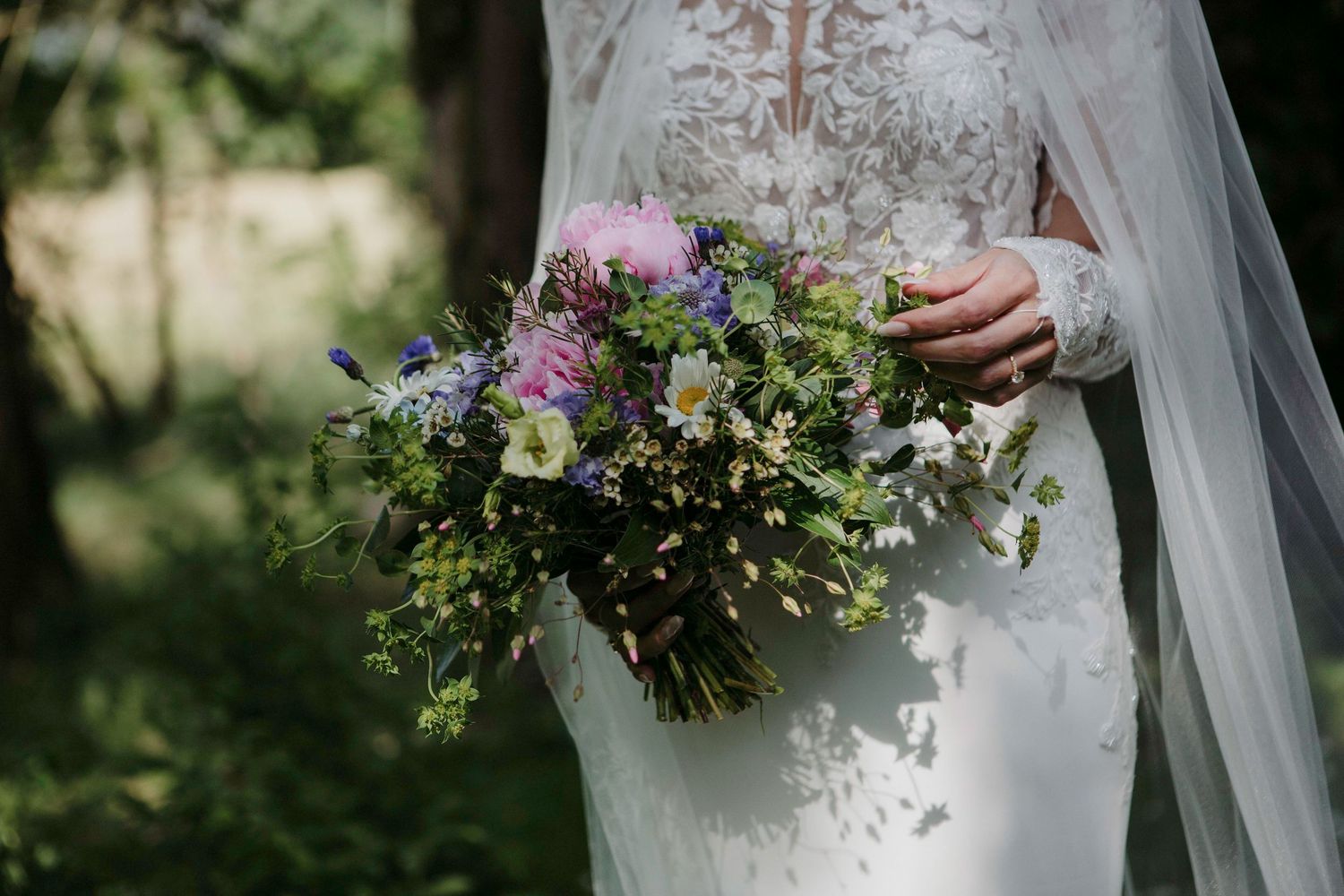 A delicate bridal bouquet with purple and pink wildflowers held against a white lace wedding dress in natural light.