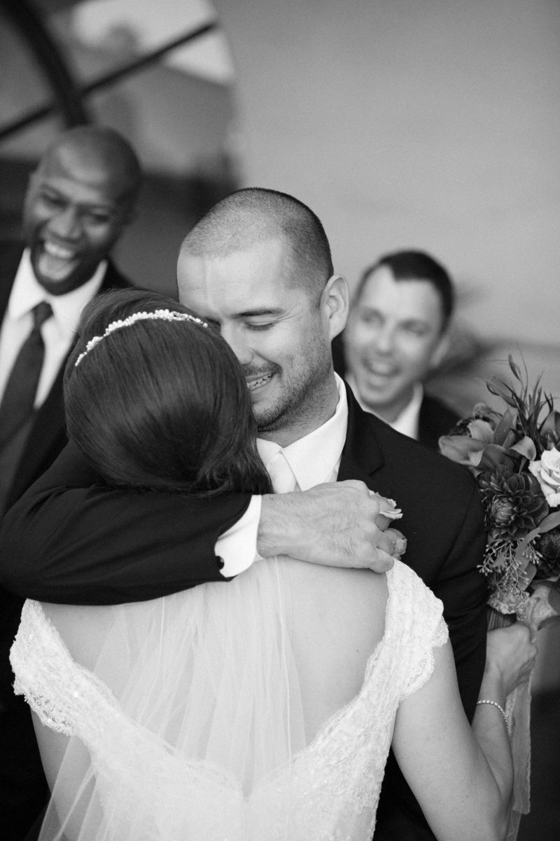 Black and white photo of a groom embracing his bride at a wedding ceremony with smiling groomsmen in the background.