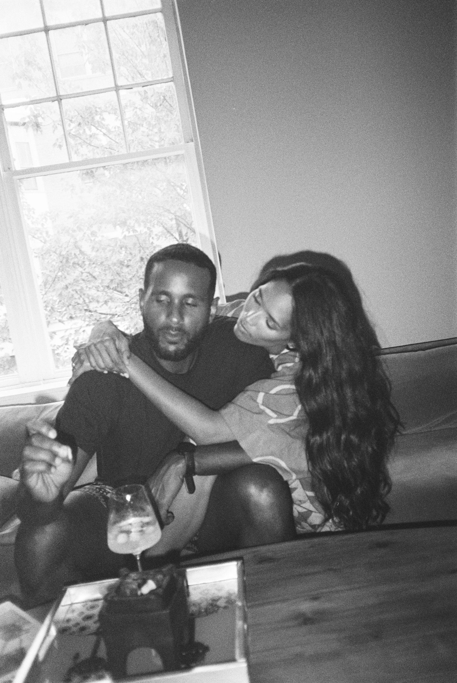 A couple shares an intimate black and white moment on a couch by a window while holding drinks.