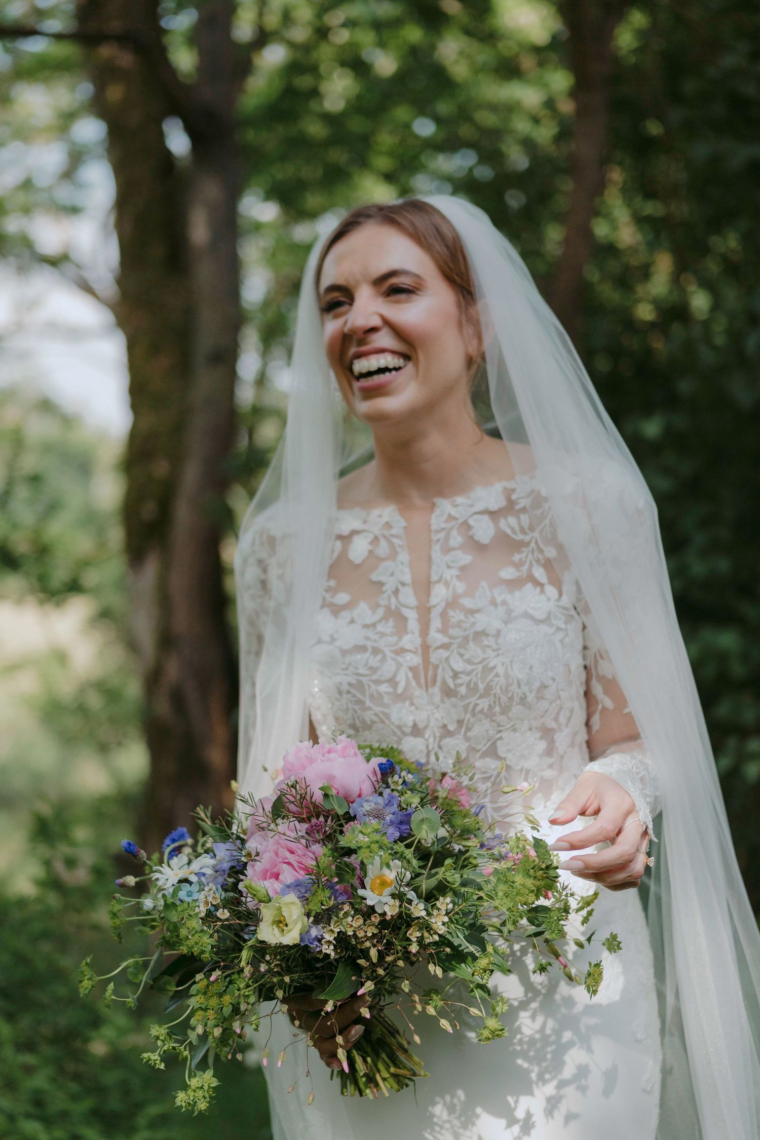 A bride in a lace wedding dress holds a colorful wildflower bouquet while smiling in a wooded outdoor setting.
