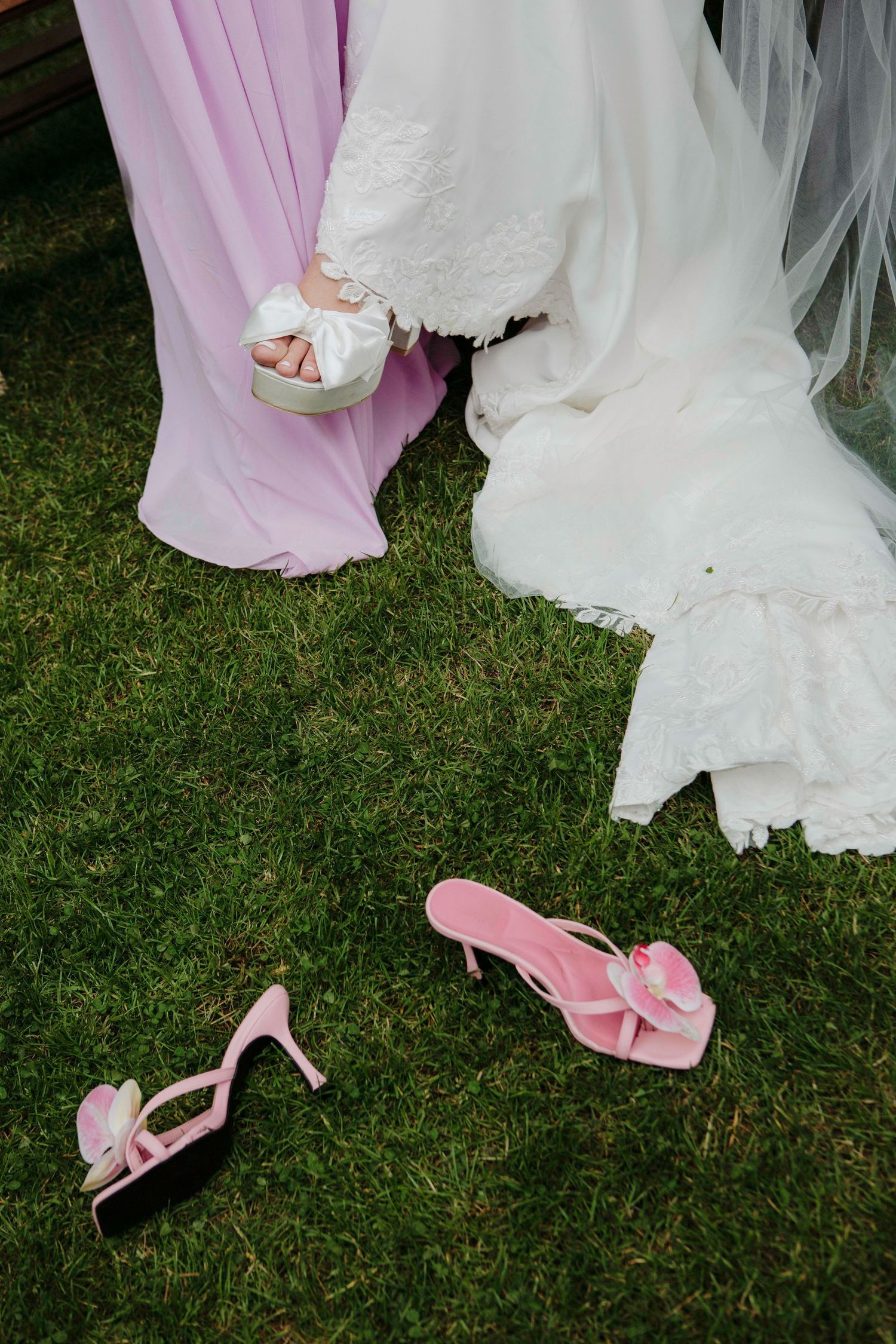 Pink and white wedding dresses with matching pink heels are arranged artistically on green grass during an outdoor celebration.