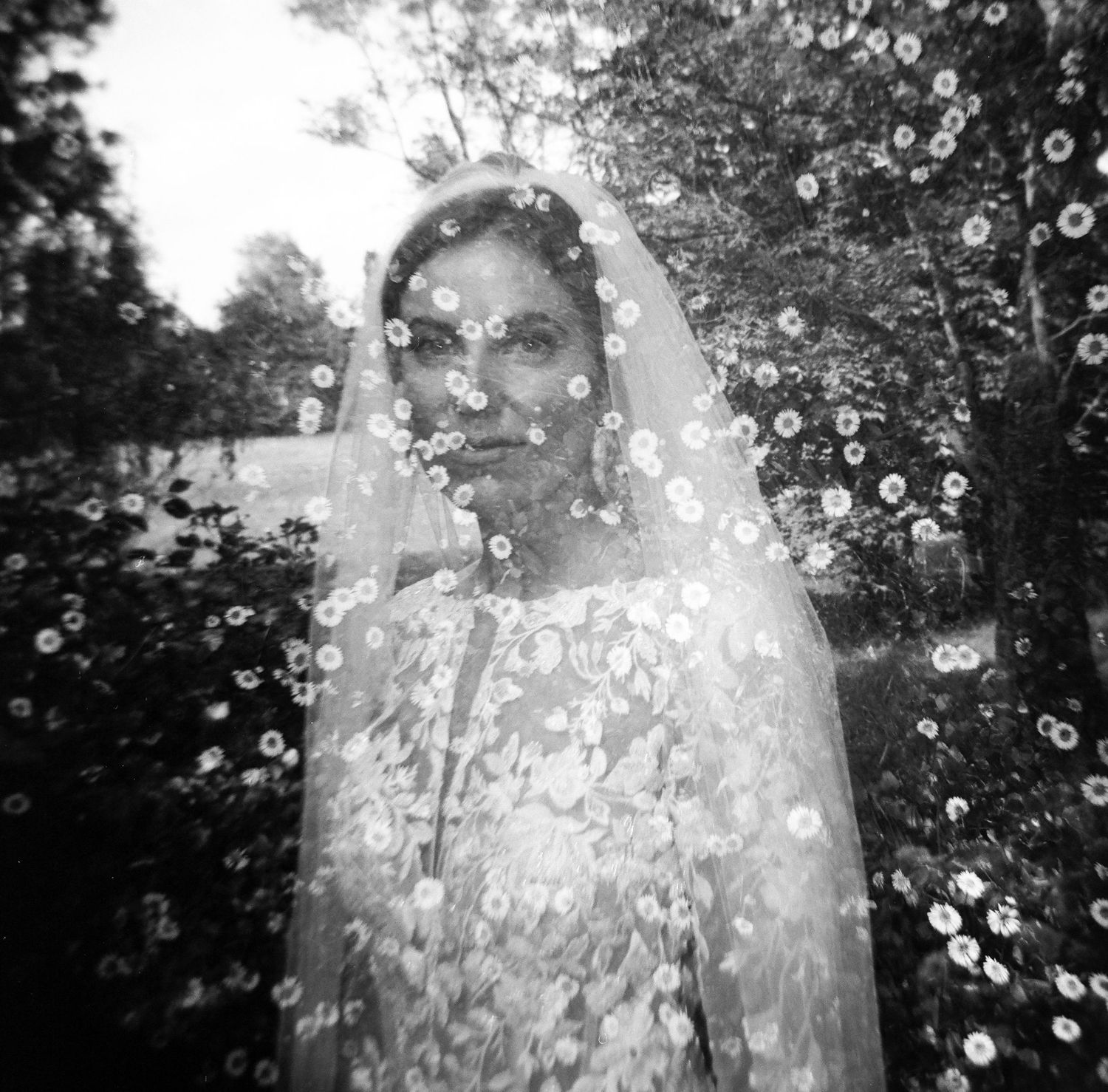 Black and white photo of a bride in floral veil and lace gown surrounded by garden greenery.