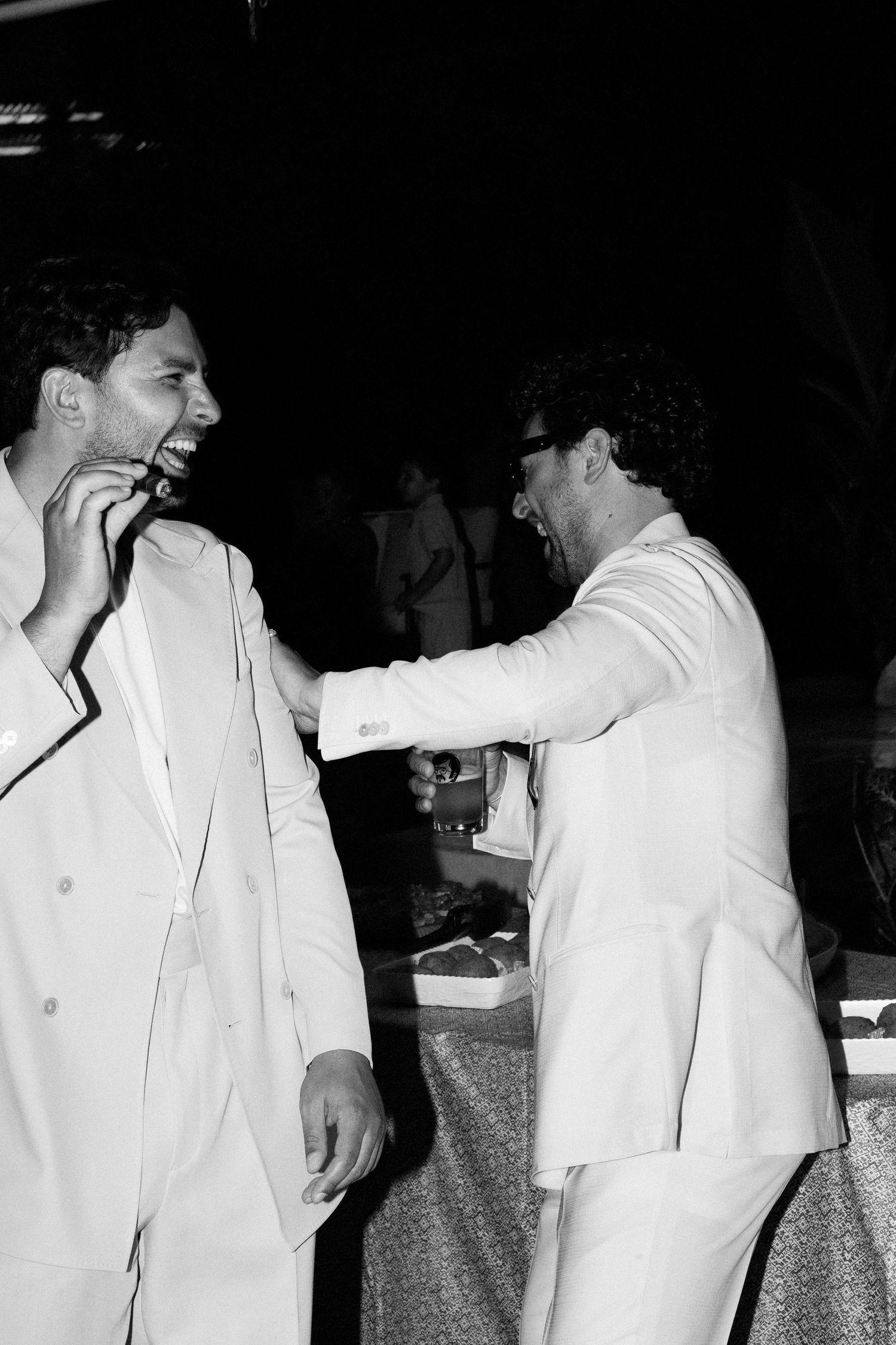 Two men in white suits laughing and sharing a joyful moment at a formal event, captured in black and white.