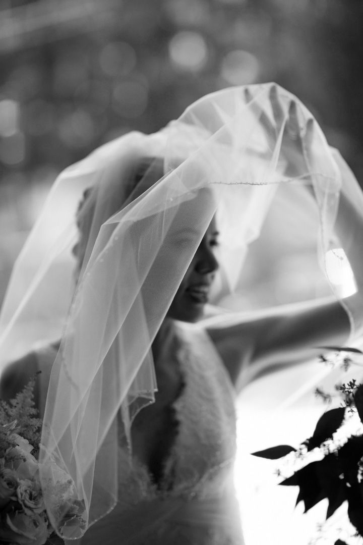 Black and white bridal portrait of a bride lifting her veil, wearing a lace wedding dress and holding a bouquet.