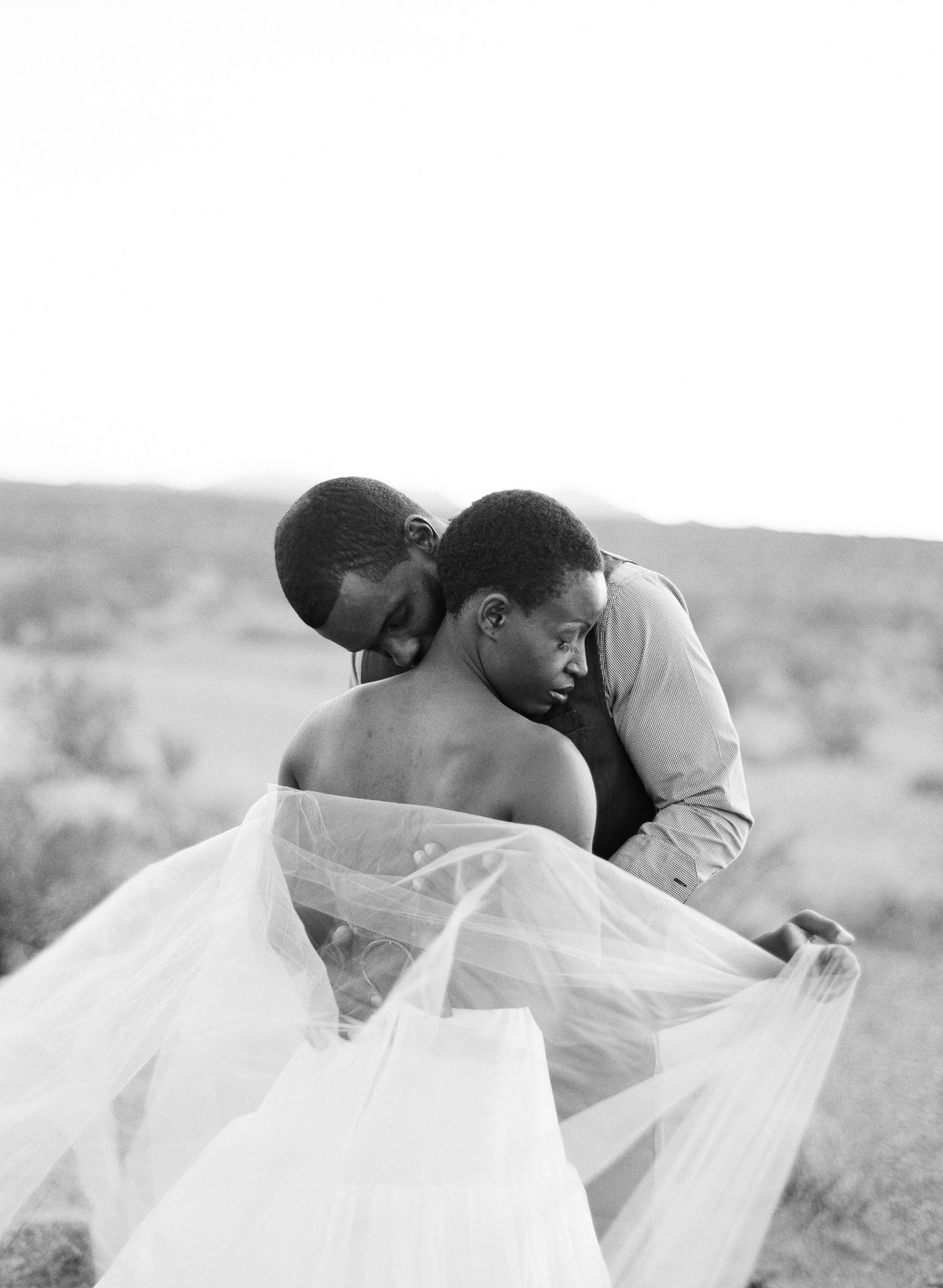 Black and white photo of two people sharing an intimate embrace outdoors, with a flowing bridal veil in the foreground.
