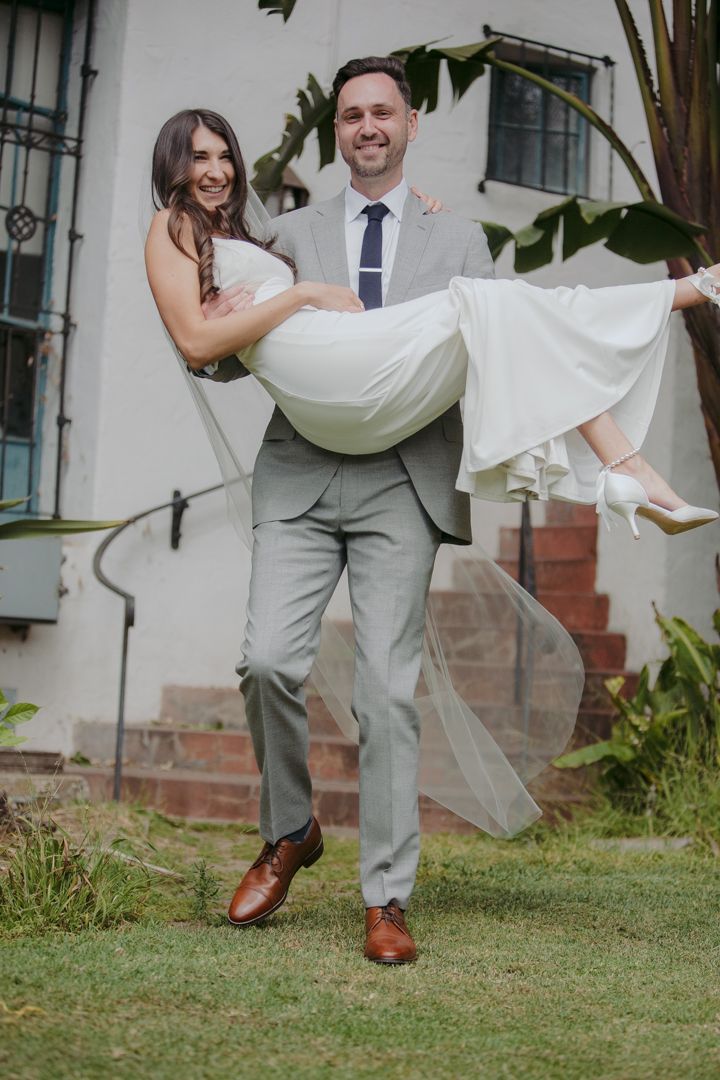 A couple in wedding attire playfully poses together on a grassy lawn outside a white building.