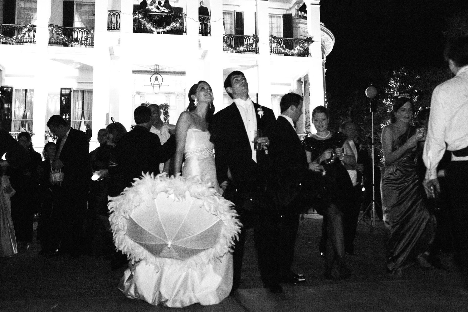 Black and white photo of a bride and groom at an outdoor evening wedding reception with guests mingling in background.