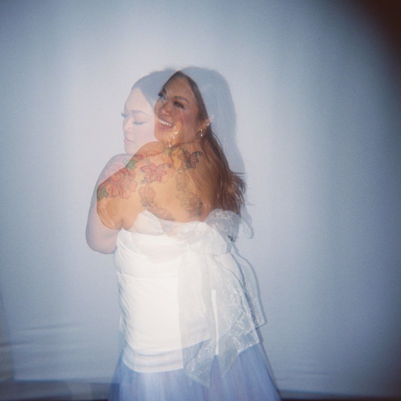 A smiling woman with tattoos wearing a white dress poses gracefully against a light background.