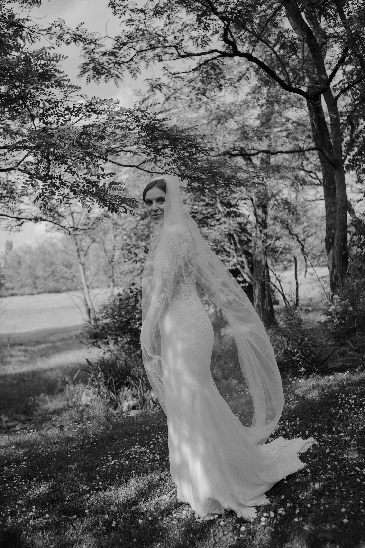Black and white photo of a bride in a flowing lace veil and wedding gown standing among trees outdoors.