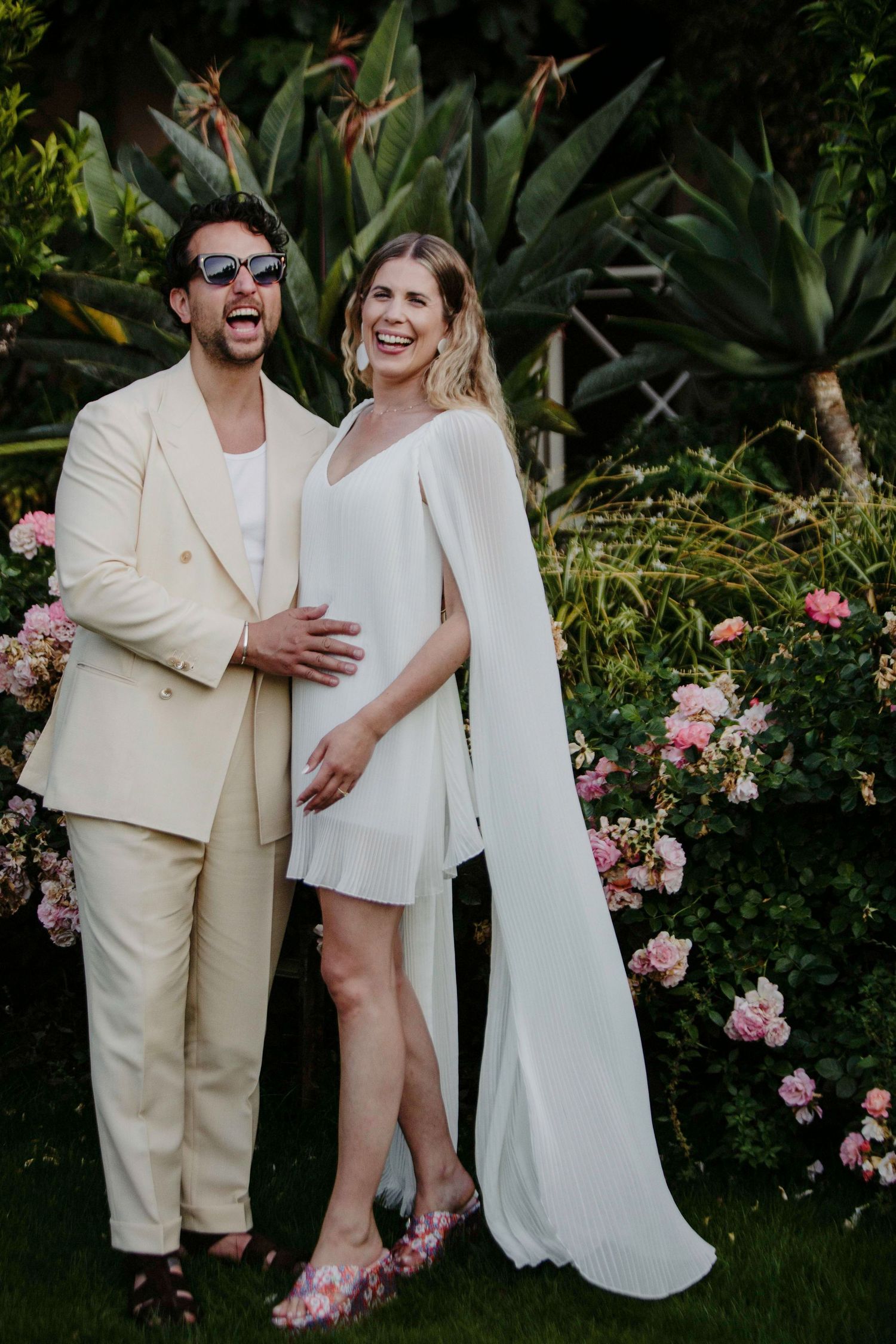 A couple in cream-colored outfits pose together in a garden with blooming pink roses.