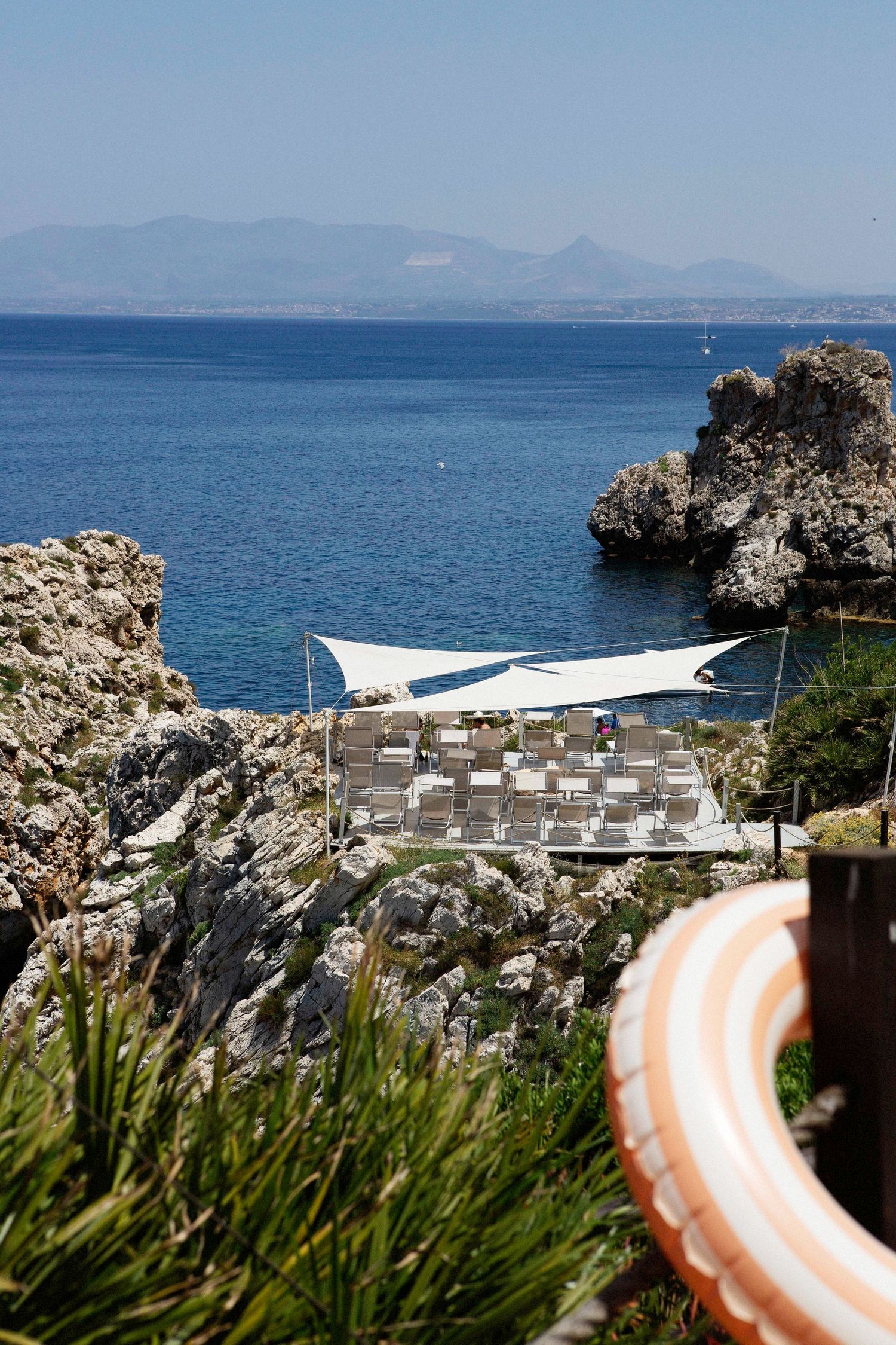 Scenic coastal view of a Mediterranean beach with white sun shade and rocky cliffs overlooking blue waters.