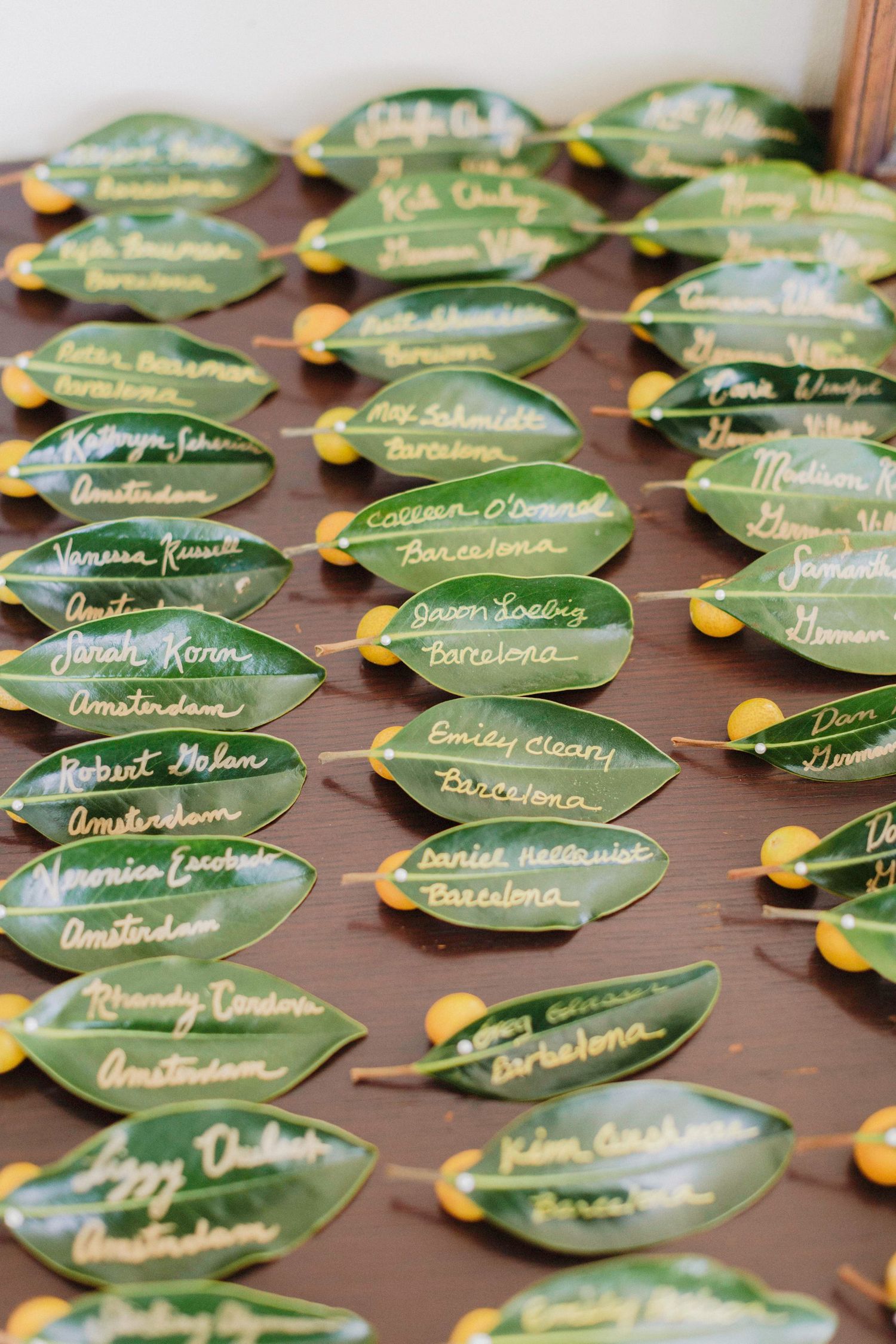 Green leaf place cards with gold calligraphy names and yellow citrus accents arranged on a wooden table for a wedding.