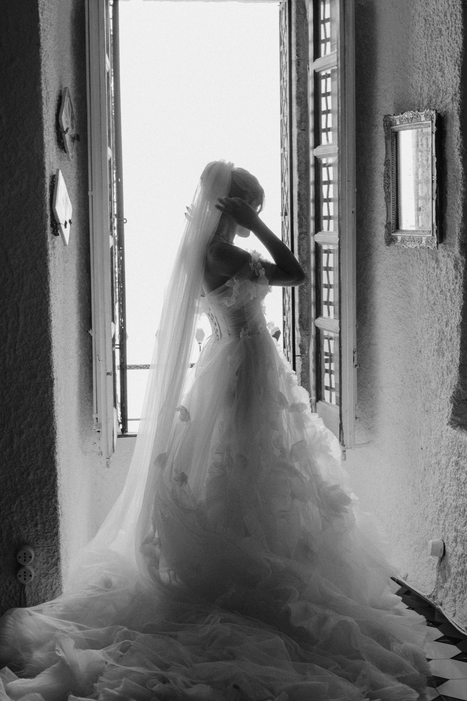 A silhouetted figure in an elegant wedding dress stands by a window in dramatic black and white lighting.