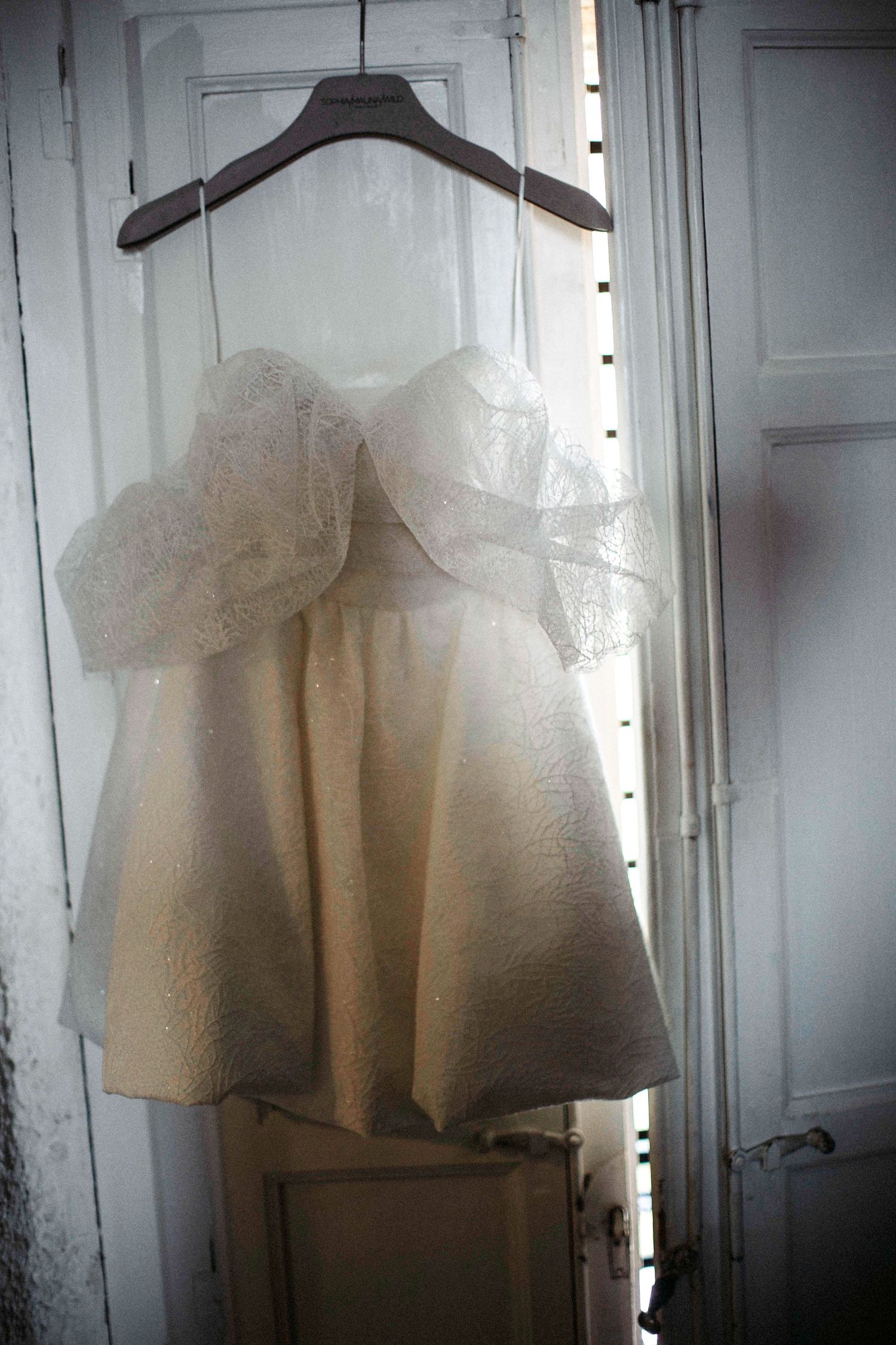 A ghostly white vintage dress hangs on a wooden hanger in front of a white door creating an ethereal atmosphere.