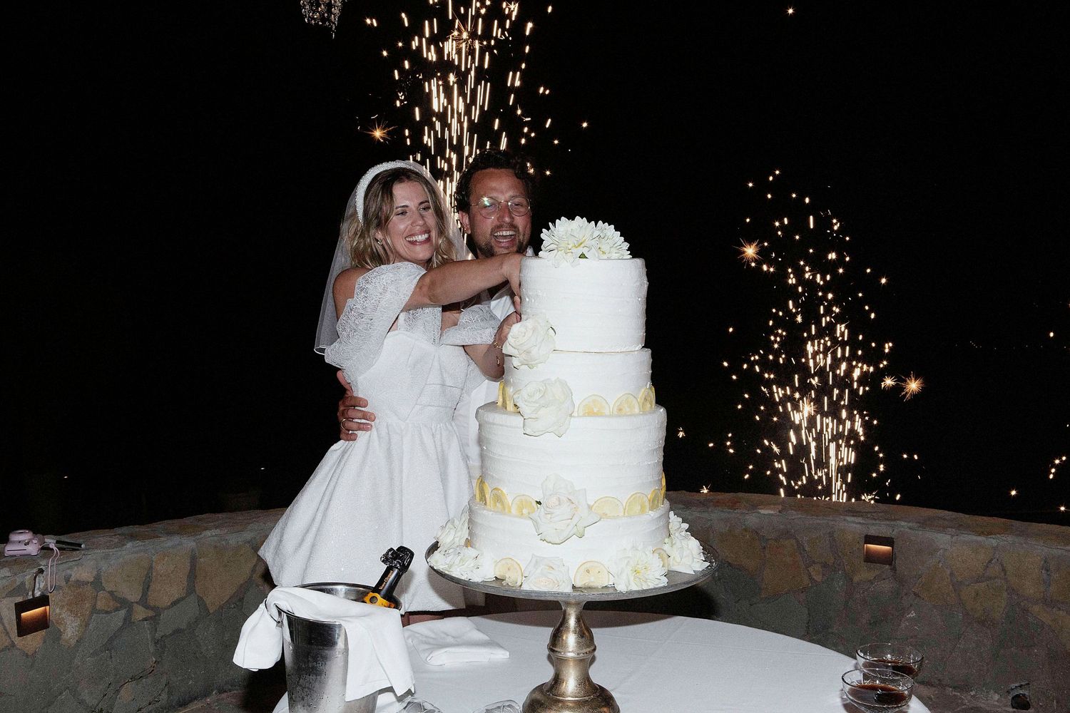A white tiered wedding cake sits on a table with sparklers creating a dramatic light display in the background.