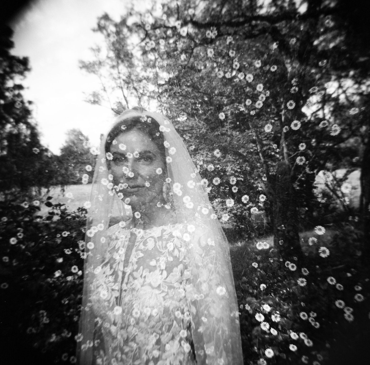 Black and white double exposure photo of a bride in veil surrounded by blooming flowers and trees.