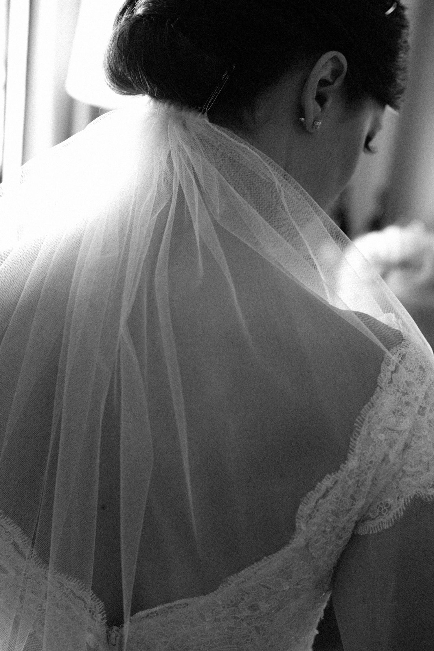 Black and white photo of a bride in a lace wedding dress with veil draped over her back, viewed from behind.