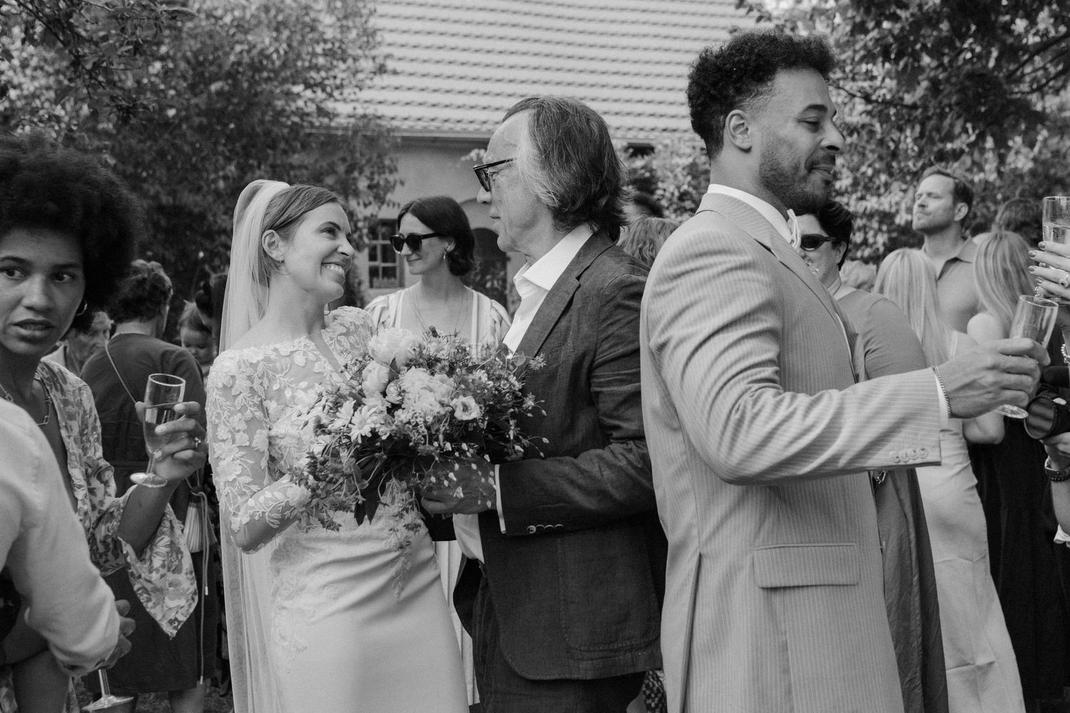 Black and white wedding photo of bride in lace gown with bouquet, groom in suit, and guests celebrating outdoors.