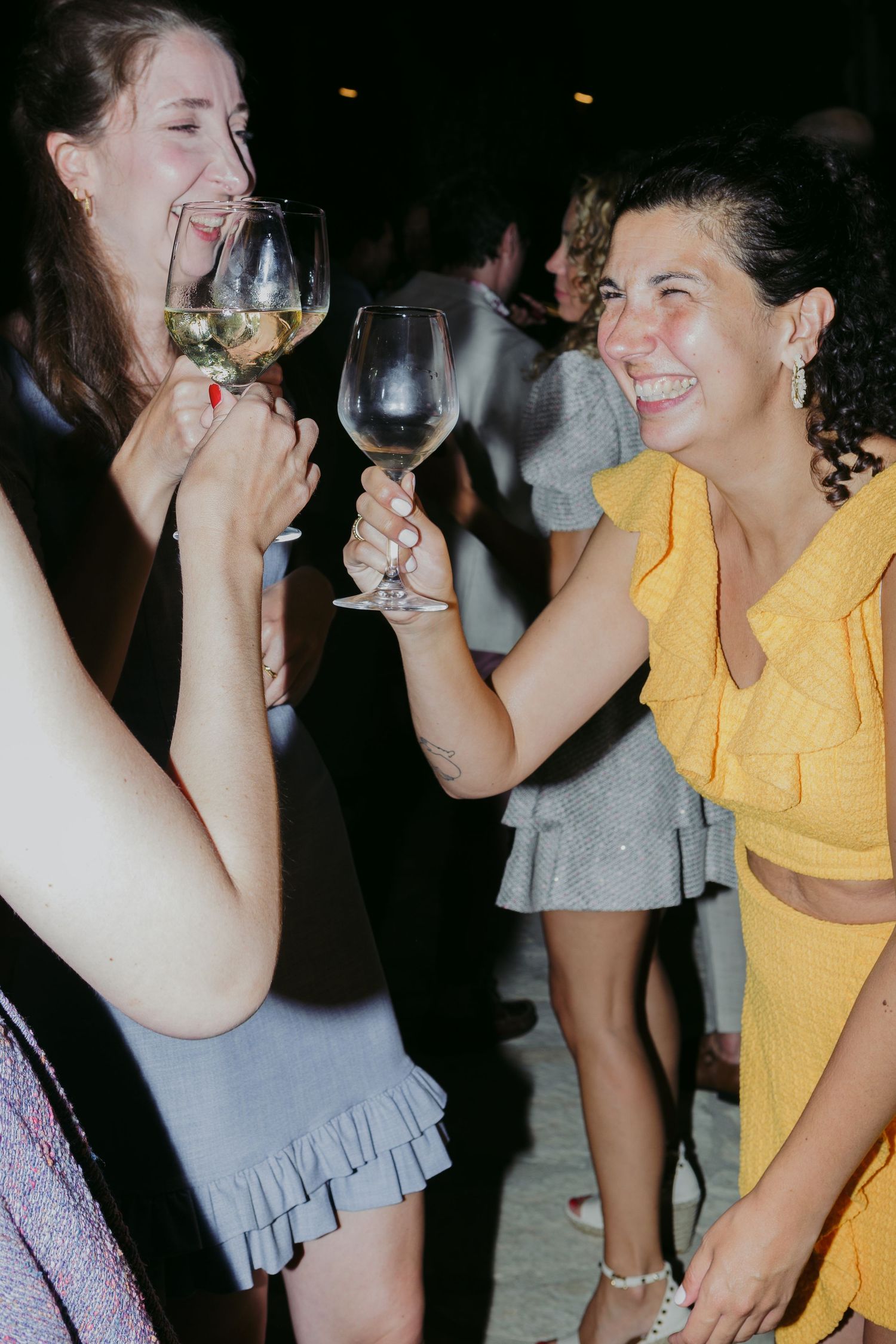 Two people in yellow and gray outfits share laughter while holding wine glasses at a nighttime gathering.