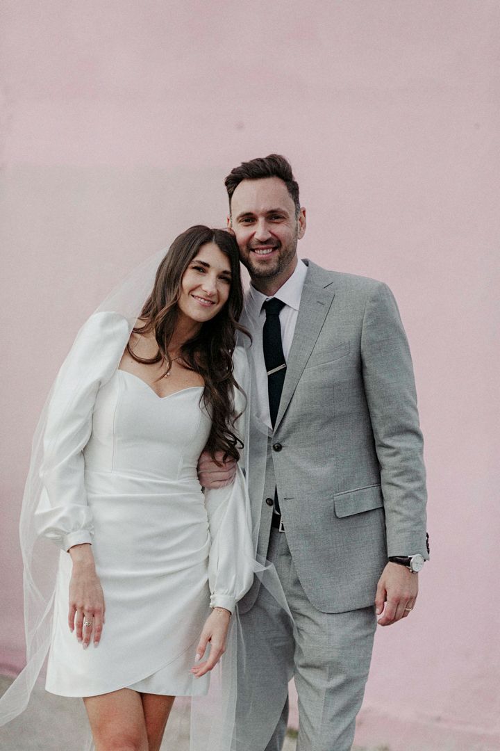 A happy couple poses together against a pink wall, the bride in a white dress and groom in a light gray suit.