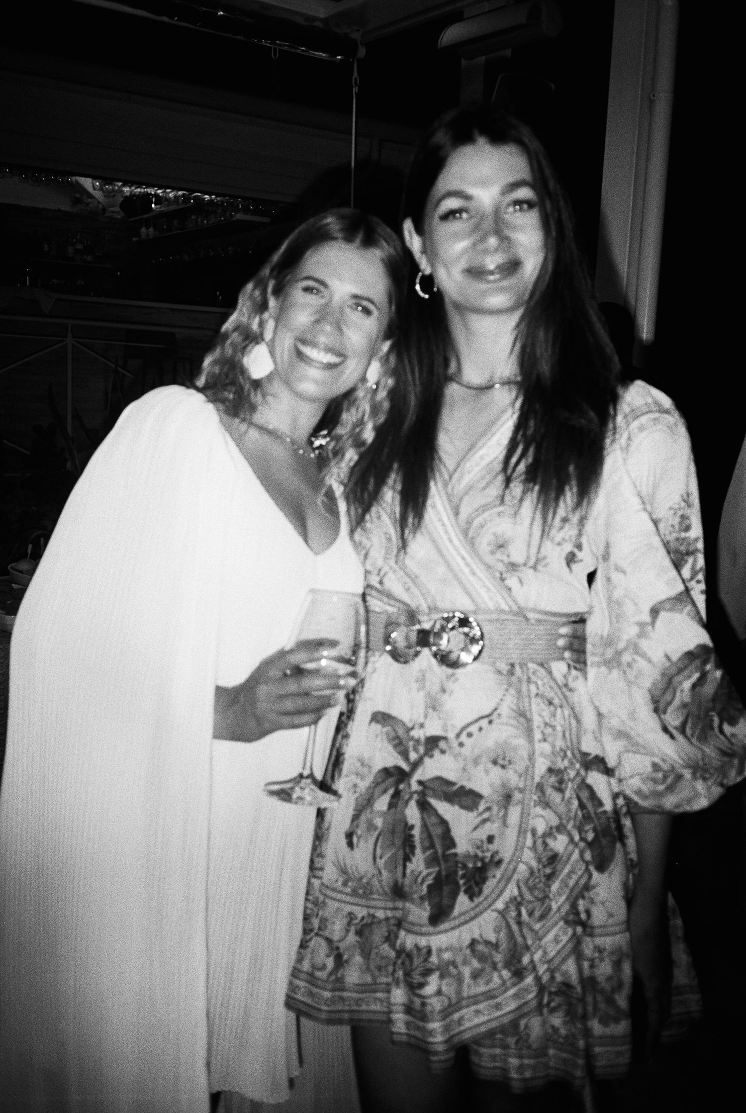 Two women smile at a social gathering, one wearing white and one in a patterned dress, holding drinks in black and white photo.