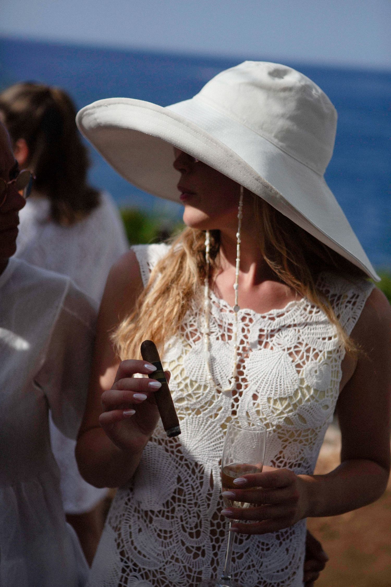 Someone in a white lace dress and wide-brimmed sun hat enjoys a sunny day by the ocean.