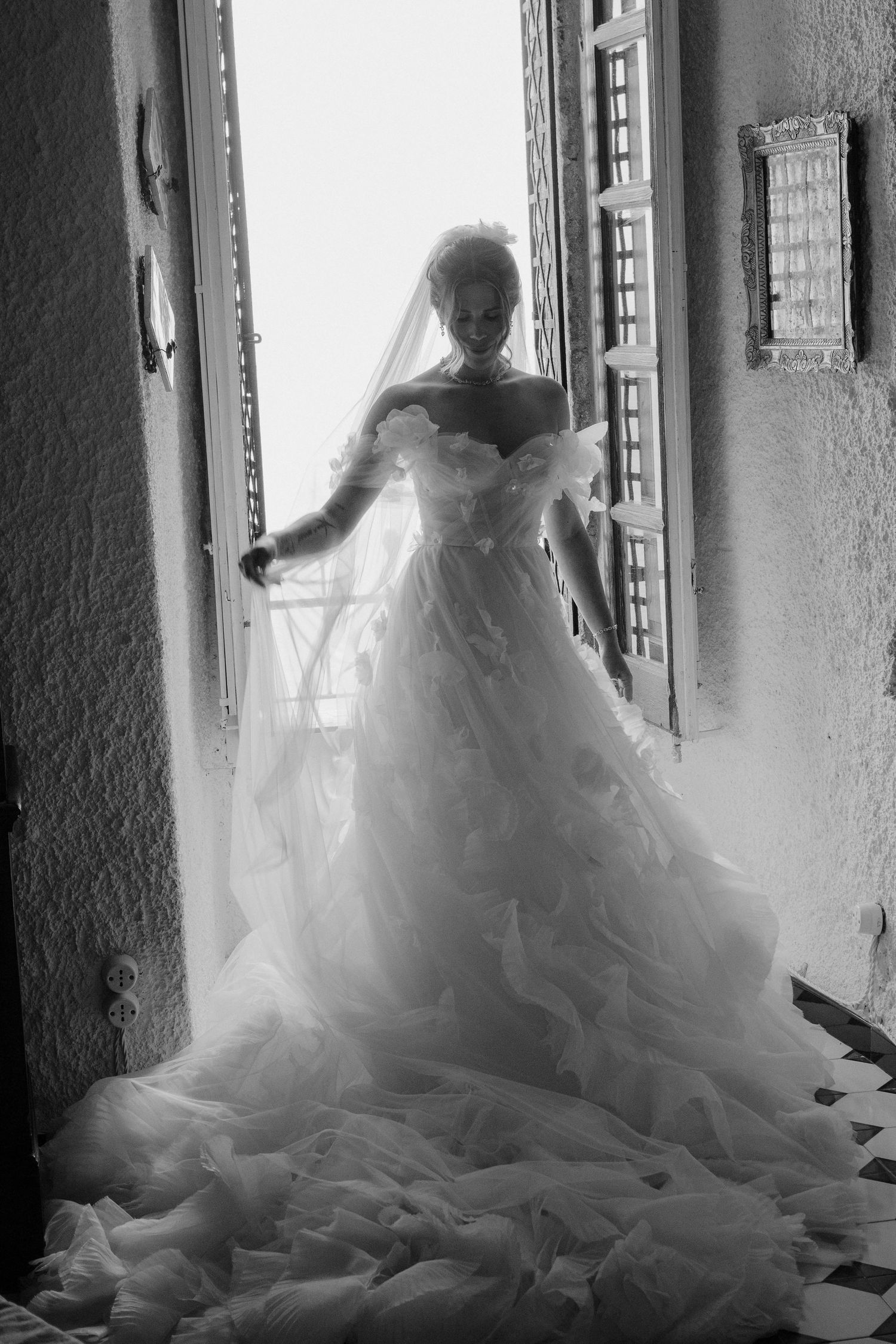 A black and white silhouette of a bride in a flowing wedding gown stands gracefully by a window in dramatic lighting.