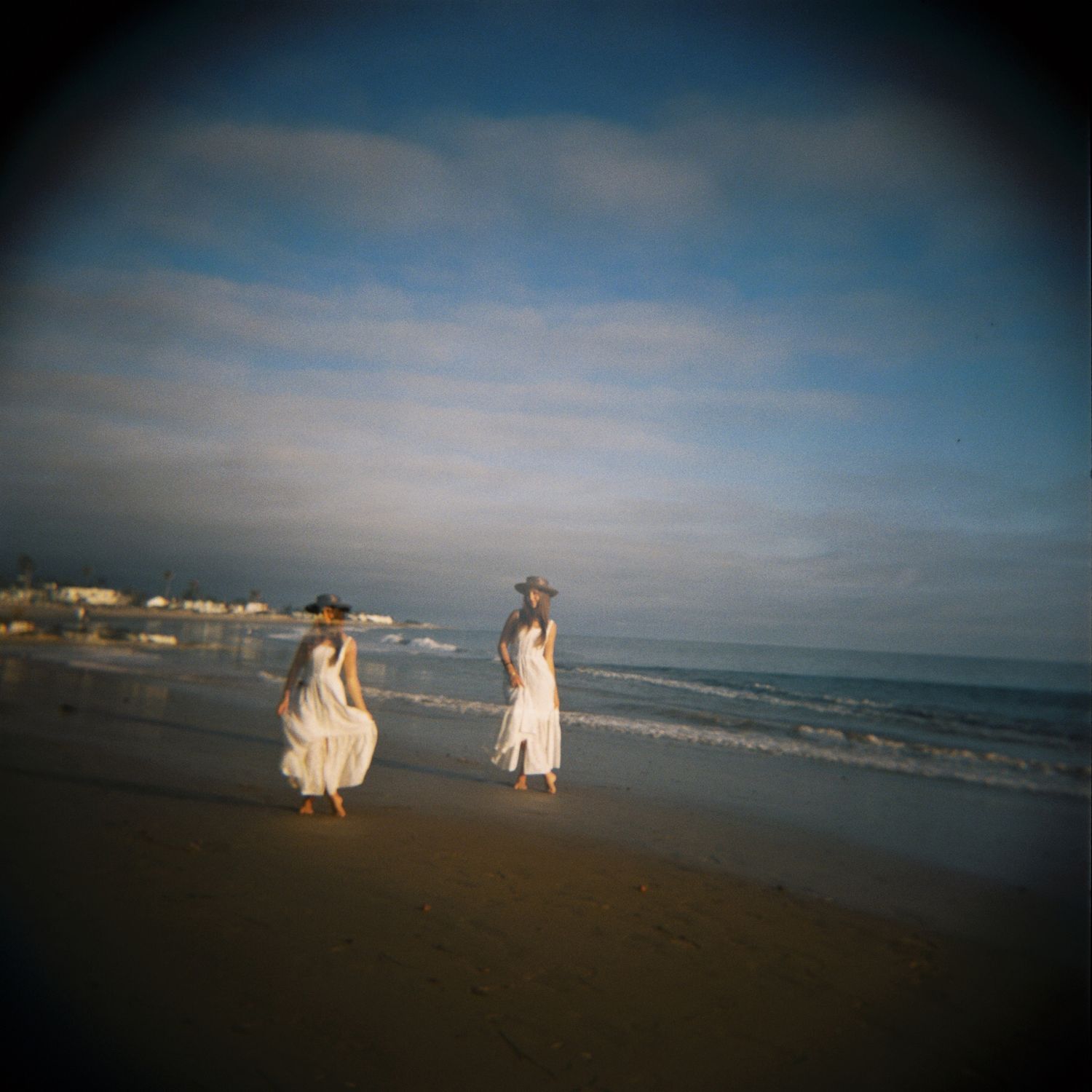 Two people in white dresses walk along a sandy beach at sunset with ocean waves and cloudy sky in the background.