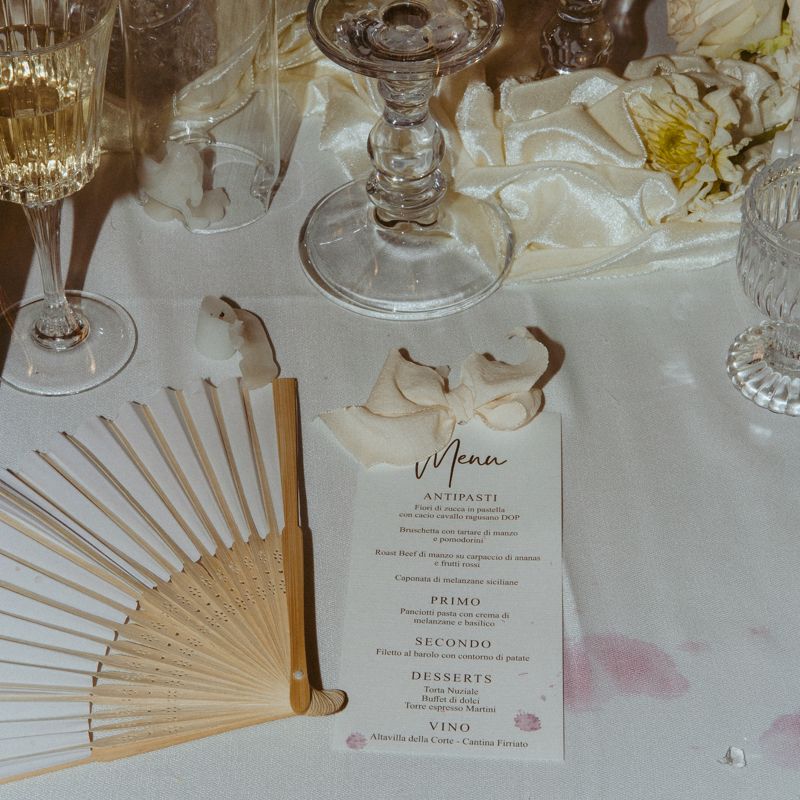 Elegant wedding table setting with folded fan, crystal glassware, white flowers and watercolor menu card on white tablecloth.